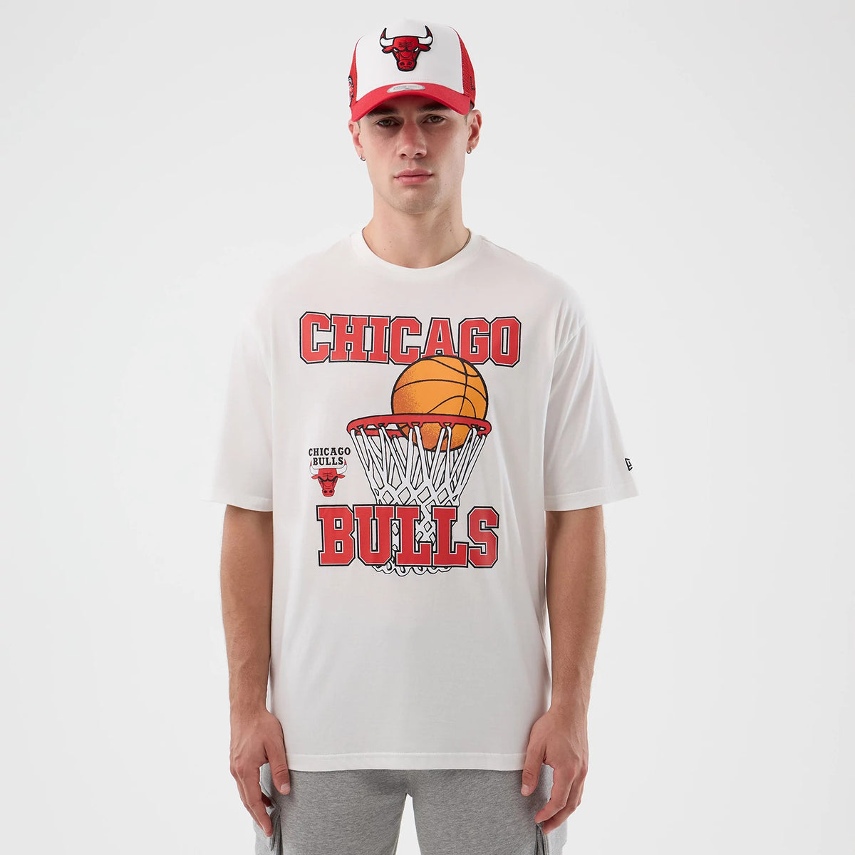 New Era Chicago Bulls NBA Basketball Graphic White Oversized T-Shirt, front view on a model with a red and white cap, featuring red Chicago Bulls lettering with a basketball and hoop graphic printed on the chest.