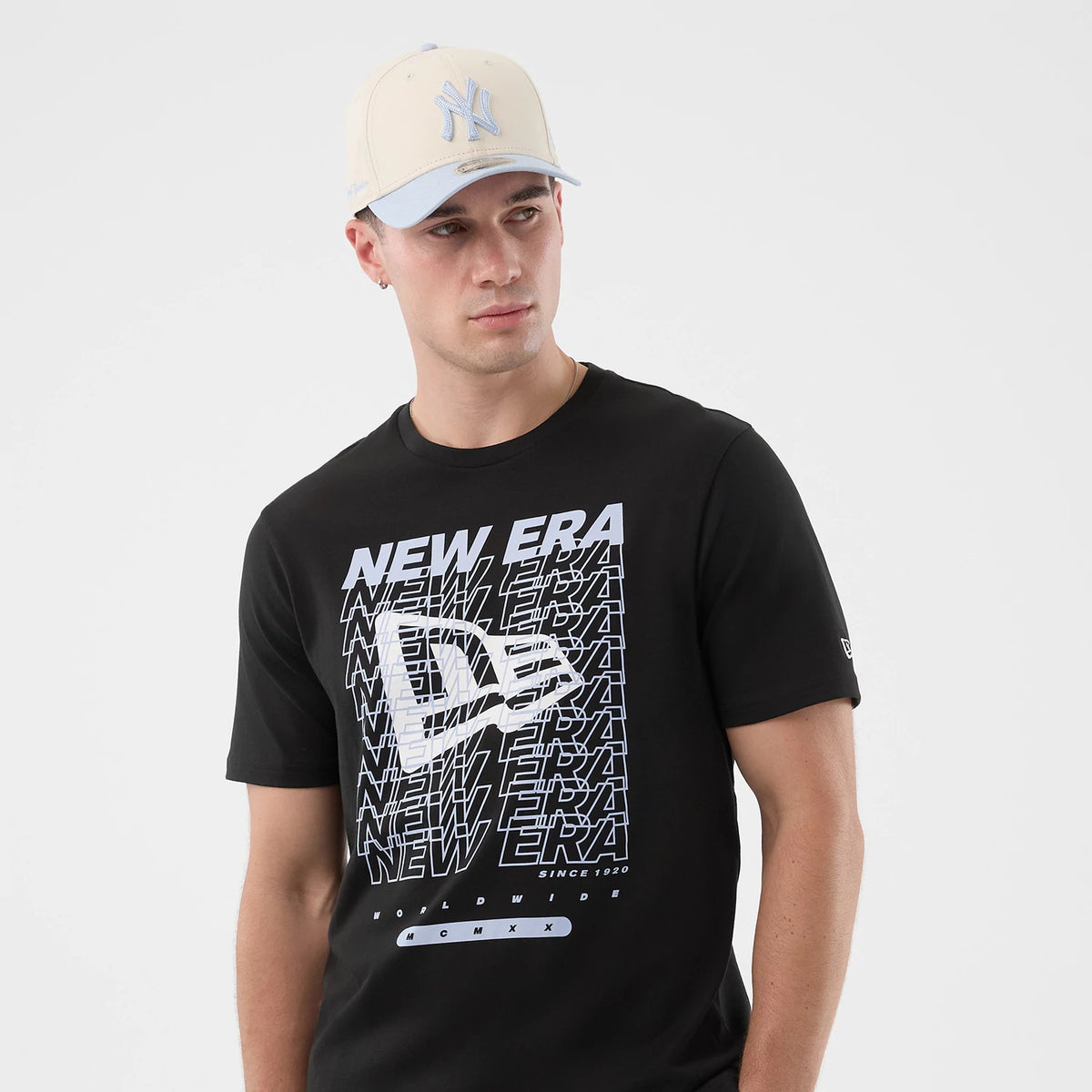 New Era Logo Graphic Black T-Shirt, front view on a model wearing a cream and blue cap with an embroidered logo, displaying a large white graphic featuring the New Era logo and layered text across the chest, and a small printed logo on the sleeve.
