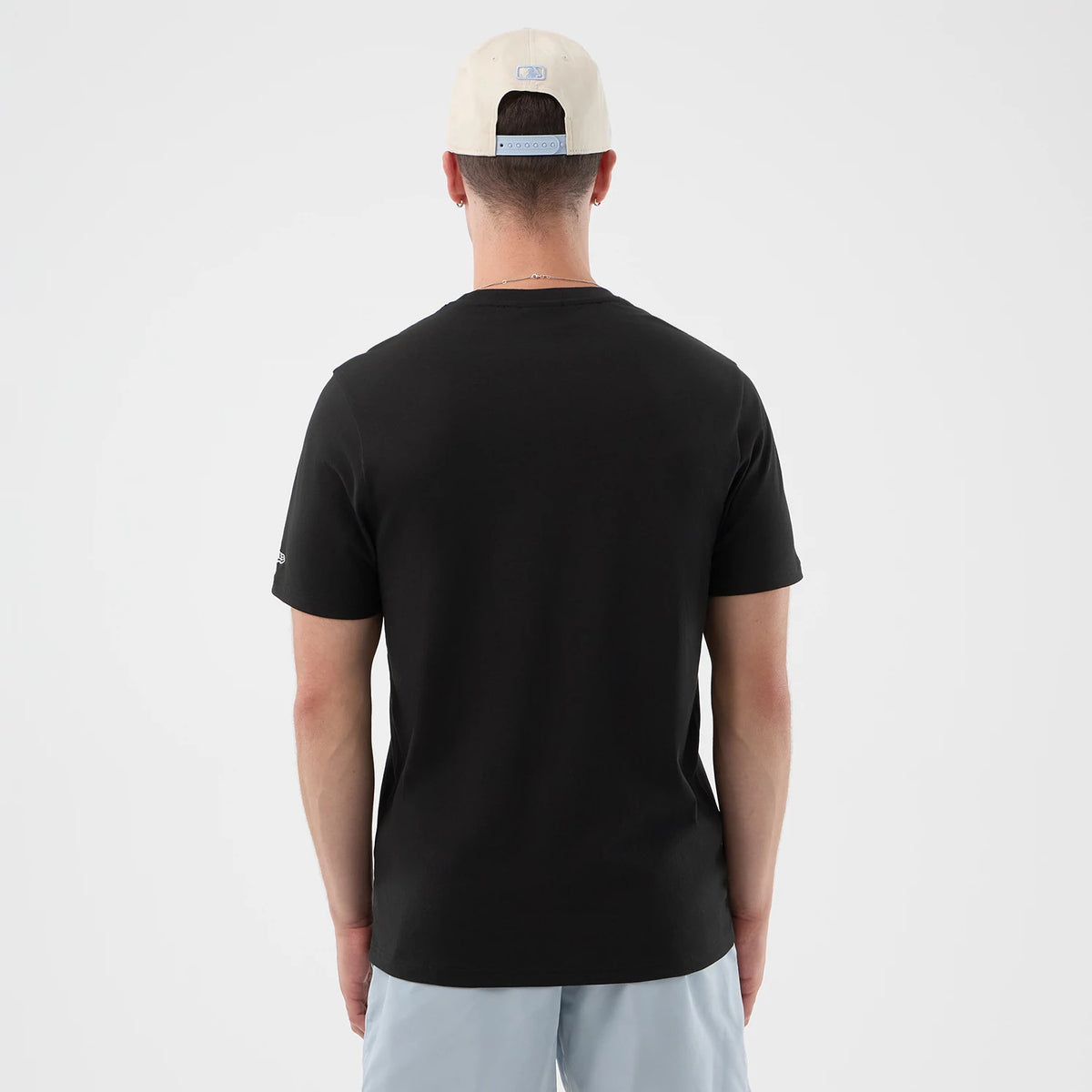 New Era Logo Graphic Black T-Shirt, back view on a model wearing light blue shorts and a cream cap, showing a plain black back panel with short sleeves and an embroidered New Era logo on the left sleeve near the cuff.