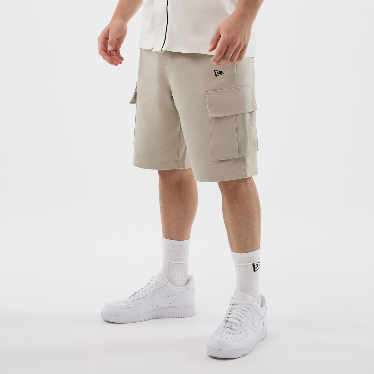 New Era Light Beige Cargo Shorts, on-model view showing two large flap pockets on the right leg and a smaller pocket on the left, a black embroidered New Era logo near the pocket, styled with a white shirt and white trainers.