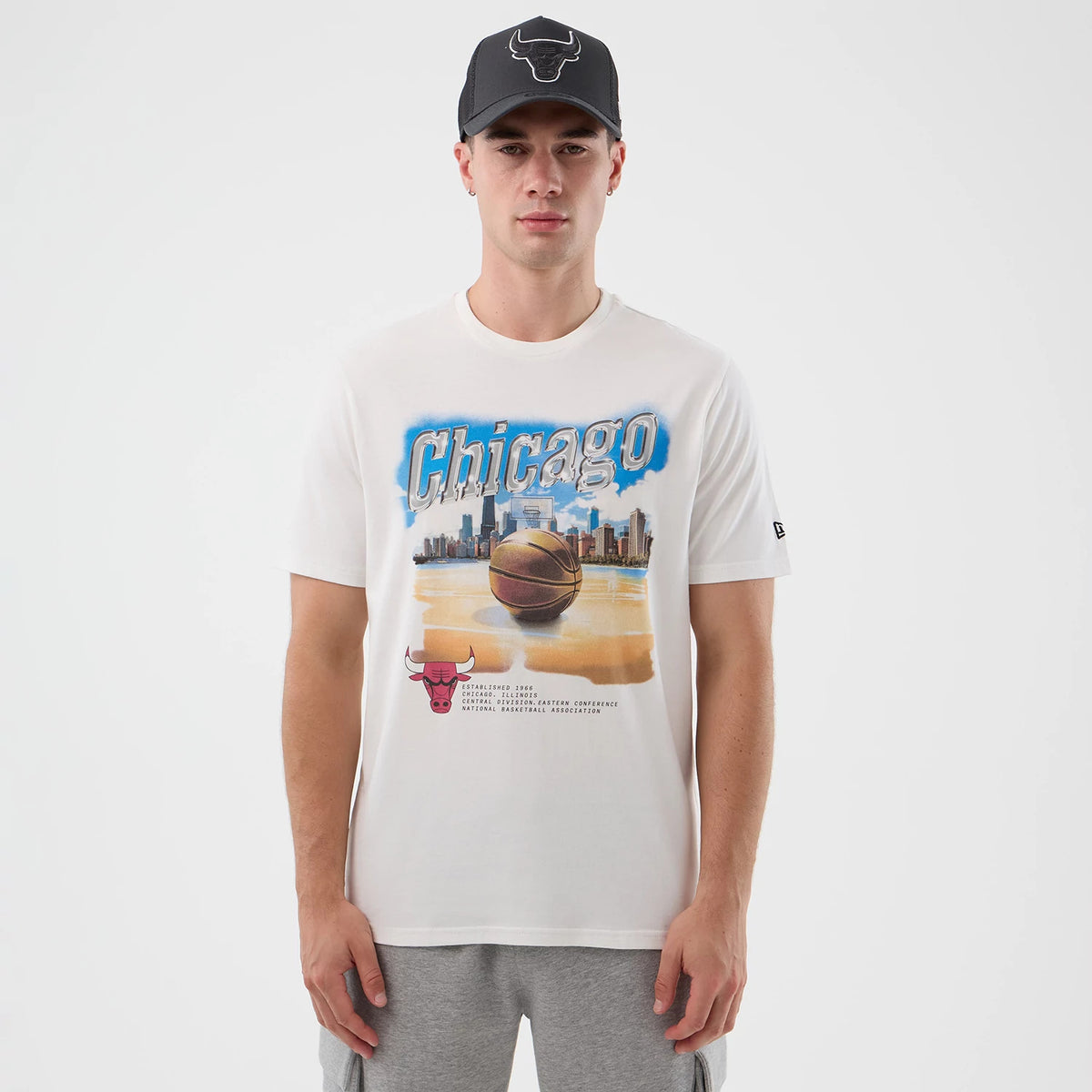 New Era Chicago Bulls NBA Beach Graphic White T-Shirt, front view on-model displaying a beach basketball court graphic with Chicago skyline, large Bulls logo at the bottom, and a black embroidered New Era logo on the sleeve.