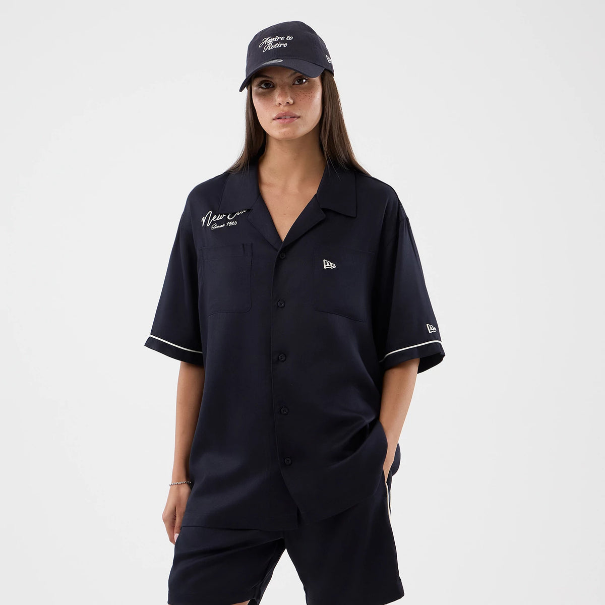New Era Graphic Revere Navy Short Sleeve Shirt, front view on a model with a matching navy cap, featuring the embroidered 'New Era Since 1920' text on the left chest, New Era flag logo on the chest pocket, and white piping on the sleeves.