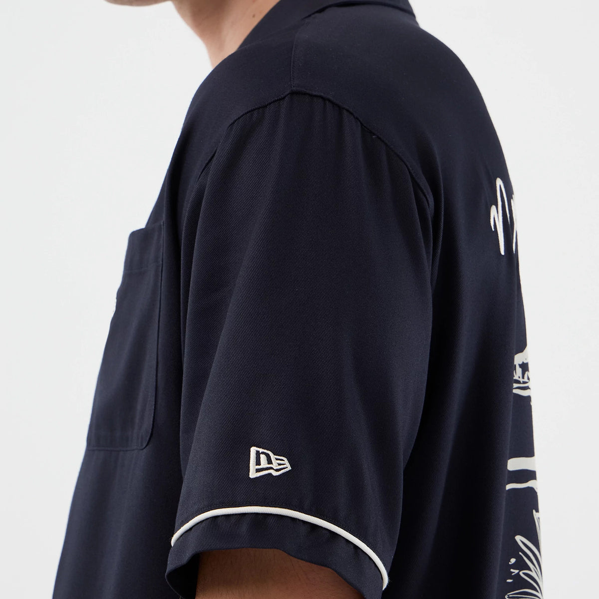 New Era Graphic Revere Navy Short Sleeve Shirt, side view on a model showing the embroidered New Era flag logo near the cuff of the sleeve, contrasting white piping along the sleeve edge, and graphic detail on the back.