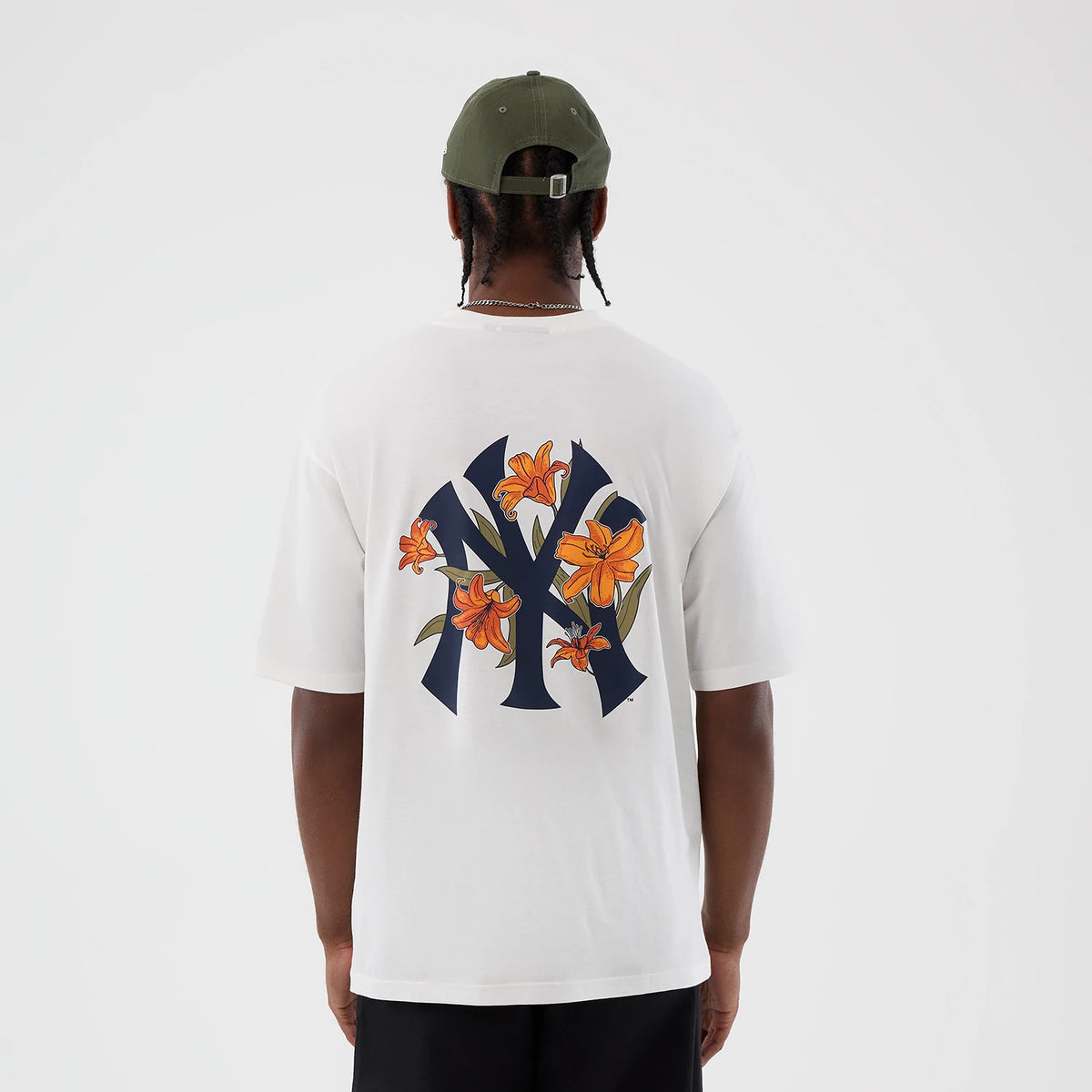 New Era New York Yankees MLB Floral White Oversized T-Shirt, back view on a model showing a large navy Yankees logo with orange floral accents across the back, paired with a green cap and black trousers, highlighting the graphic design.