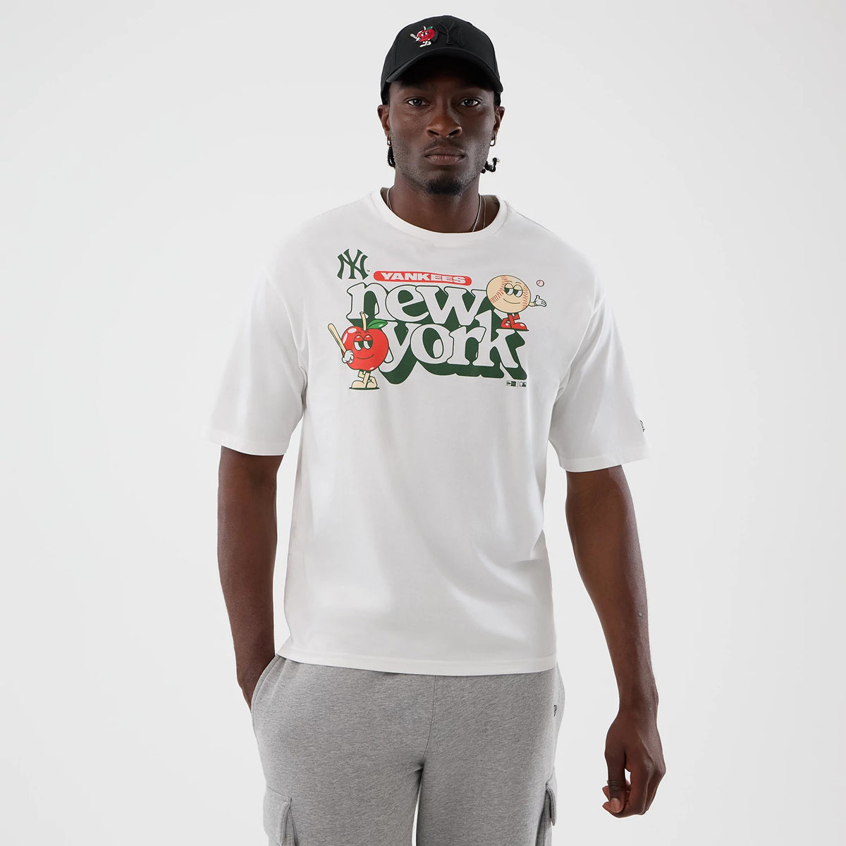 New Era New York Yankees MLB Icon Graphic White T-Shirt, front view on-model showing a green 'new york' text with cartoon graphics above, a black embroidered Yankees logo on the upper left chest, and paired with grey cargo shorts.