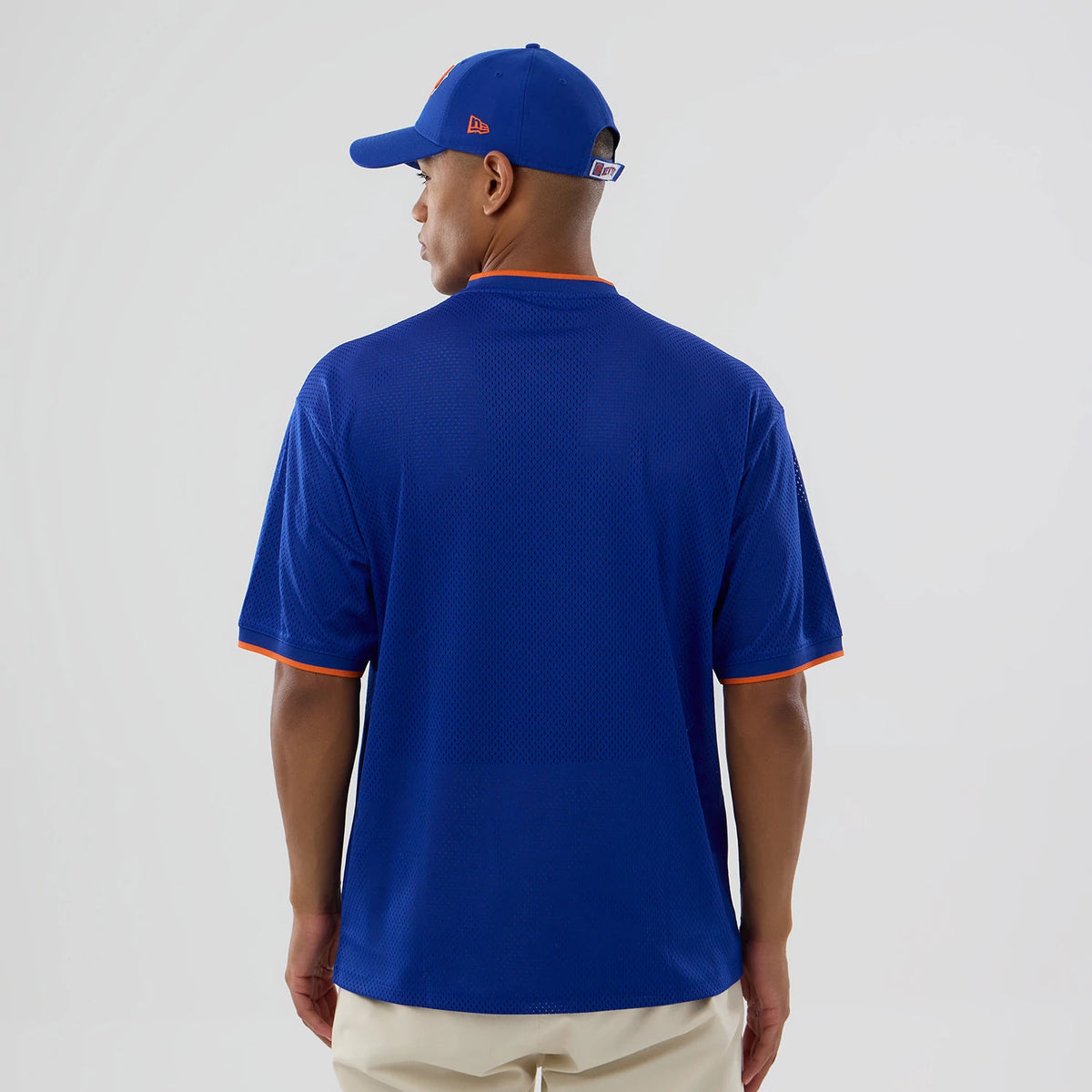 New Era New York Knicks NBA Mesh Blue Oversized T-Shirt, back view on a model wearing a matching blue cap, showing the plain mesh fabric with orange trim on the sleeves and neck, emphasising the relaxed fit and clean design.
