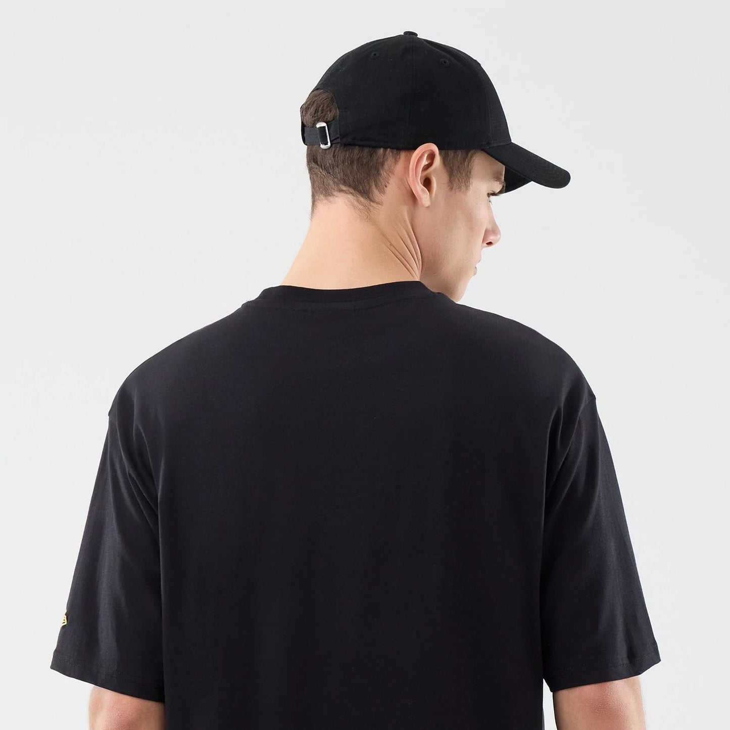 New Era Sticker Dubai Black Oversized T-Shirt, back view on a model featuring a plain black back, short sleeves, and a small yellow embroidered New Era logo on the left sleeve, styled with a black cap adjusted with a buckle.
