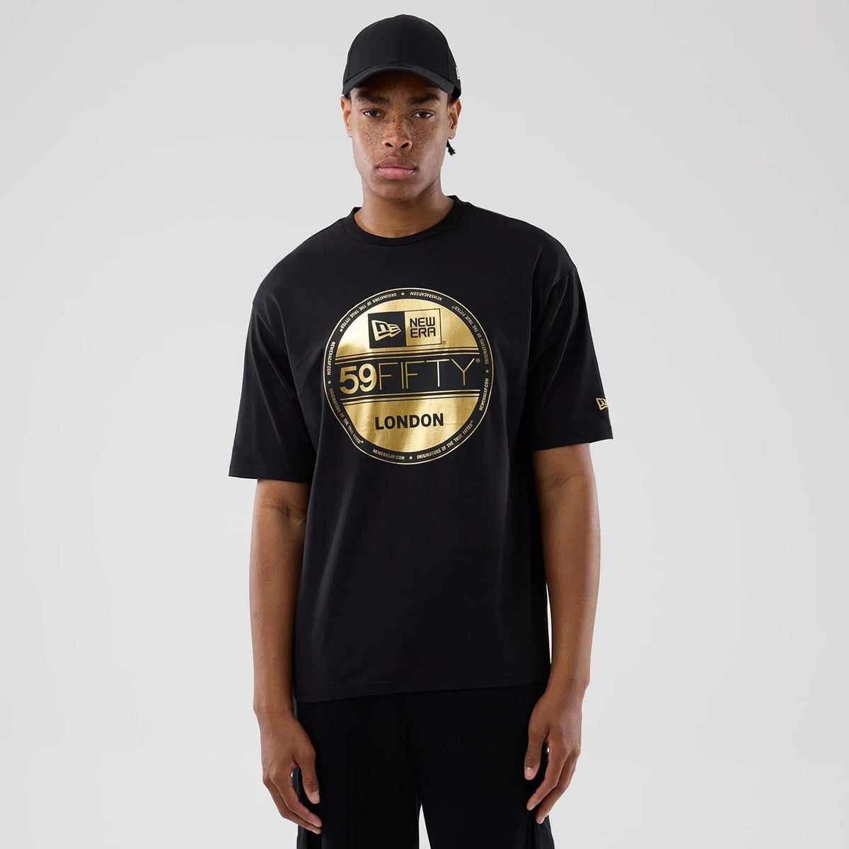 New Era Sticker London Black Oversized T-Shirt, front view displaying a large gold printed circular graphic of the New Era 59FIFTY logo and London text on the chest, with a small gold embroidered New Era logo on the left sleeve, worn by a model.