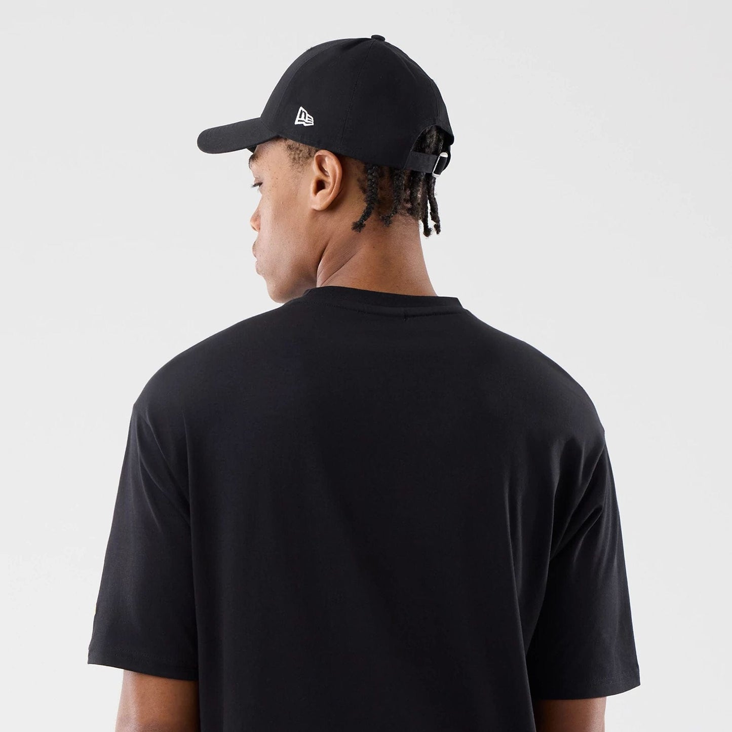 New Era Sticker London Black Oversized T-Shirt, back view on a model showing the plain black fabric with a relaxed fit, short sleeves, and a small New Era logo embroidered on the side of the black cap worn by the model.
