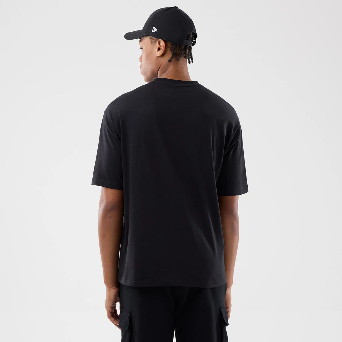 New Era Sticker London Black Oversized T-Shirt, back view on-model showcasing the plain black back with a loose fit, short sleeves, and complemented by a black cap with an embroidered New Era logo on the side of the crown.