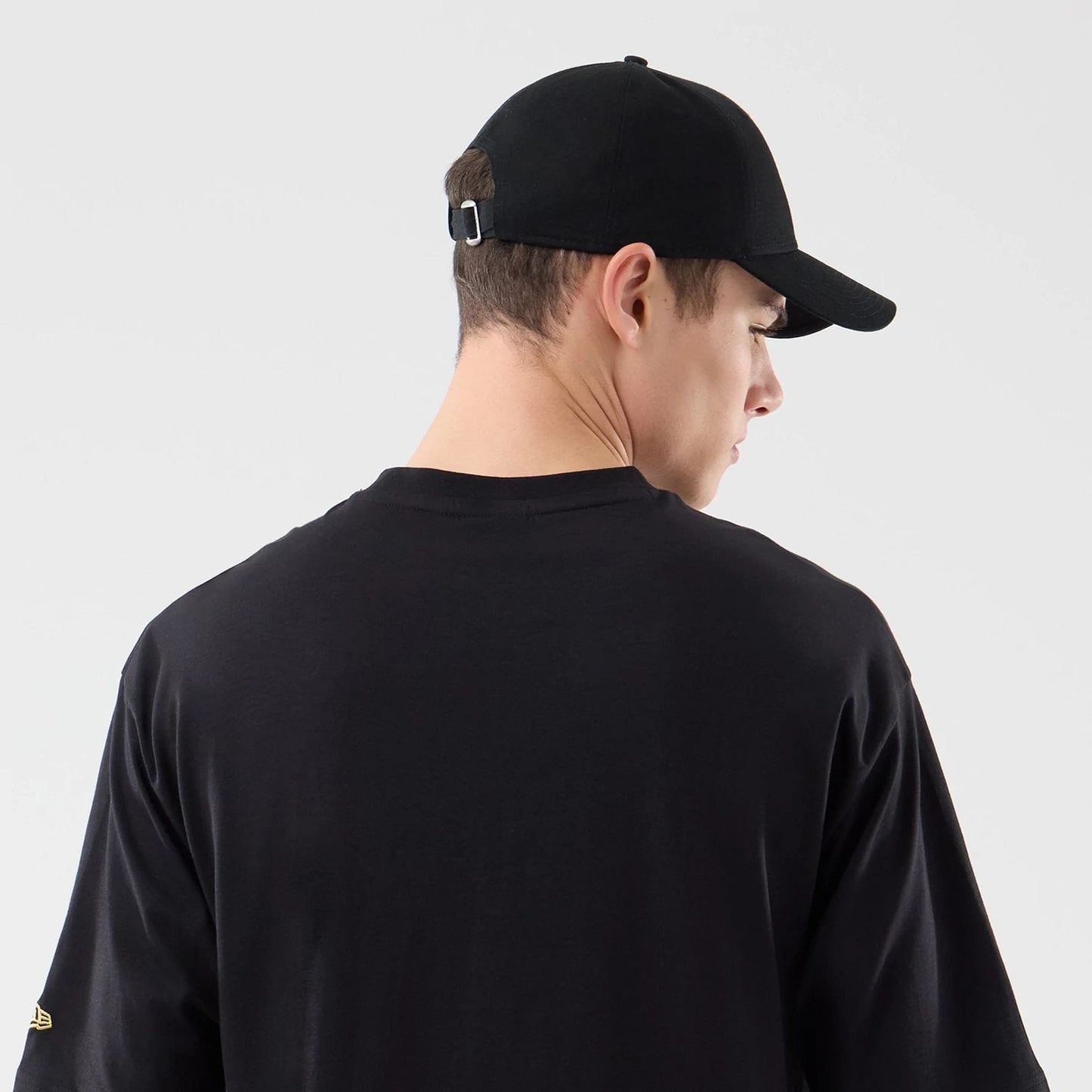 New Era Sticker Lisbon Black Oversized T-Shirt, back view on a model wearing a black cap, displaying the plain black back panel and short sleeves, with a small embroidered New Era logo on the left sleeve.