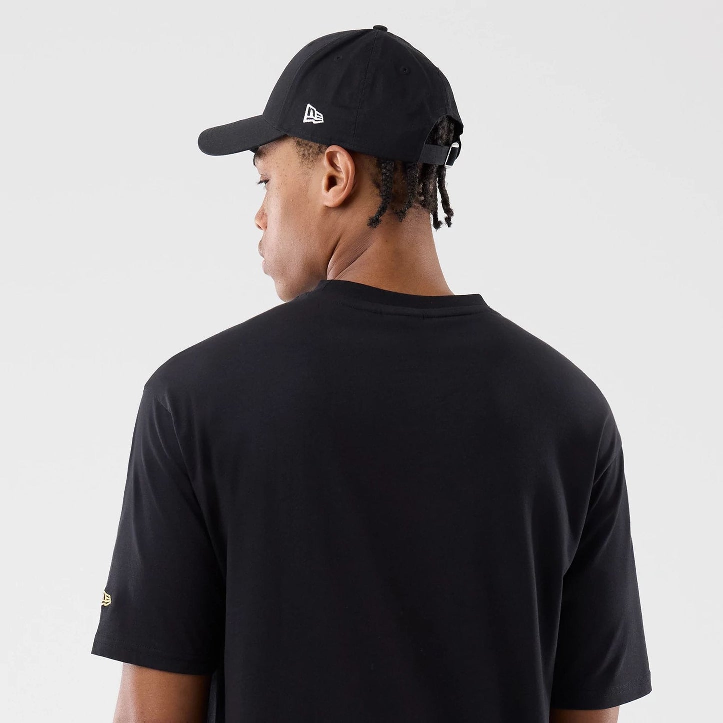 New Era Sticker Madrid Black Oversized T-Shirt, back view on a model wearing a black cap, showing plain black fabric with a small embroidered gold logo on the left sleeve and the cap having an embroidered logo on the side panel.