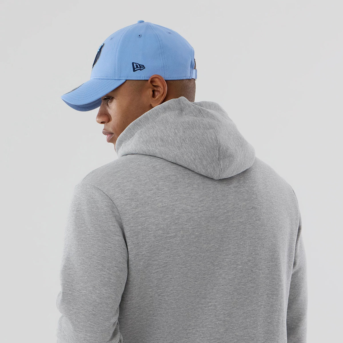 New Era Memphis Grizzlies NBA Berlin Game 2026 Grey Pullover Hoodie, three-quarter view on a model styled with a light blue cap, showing the plain grey back with the relaxed hood draped down.
