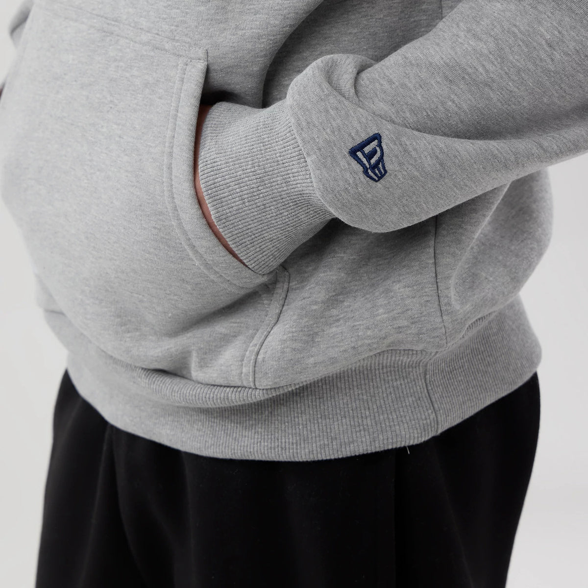 New Era Memphis Grizzlies NBA London Game 2026 Grey Pullover Hoodie, side view showing the relaxed fit with a front pouch pocket, ribbed cuffs, and an embroidered New Era logo on the sleeve, paired with black trousers for a casual look.