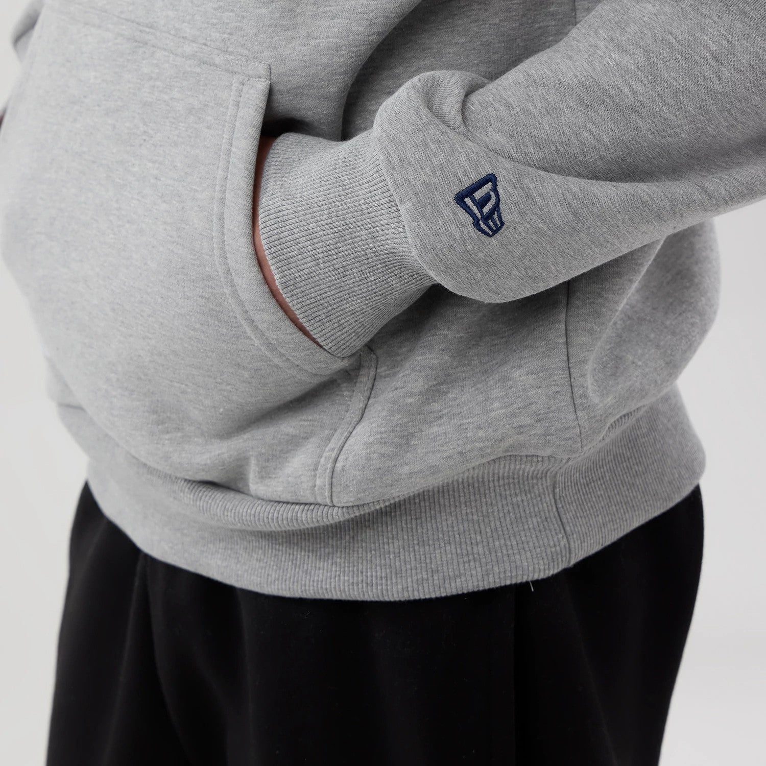New Era Memphis Grizzlies NBA London Game 2026 Grey Pullover Hoodie, side view showing the relaxed fit with a front pouch pocket, ribbed cuffs, and an embroidered New Era logo on the sleeve, paired with black trousers for a casual look.
