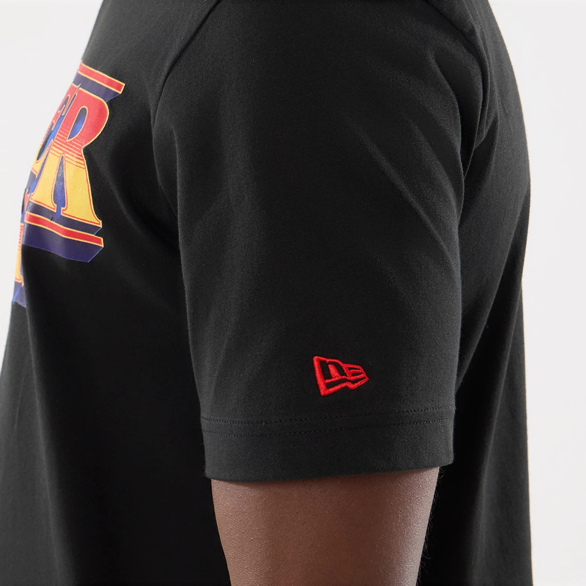 New Era Netflix Stranger Things Black T-Shirt, side view showing the red and orange logo printed on the chest, a distinct red embroidered New Era logo on the left sleeve near the hem, and a black fabric finish.