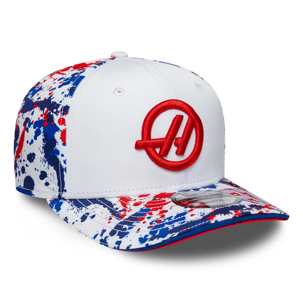 TGR Haas F1 Oliver Bearman Silverstone Race Special White New Era 9SEVENTY Stretch Snap Cap, three-quarter view showing a red raised embroidered logo on the white front panel, splatter print pattern in red and blue.
