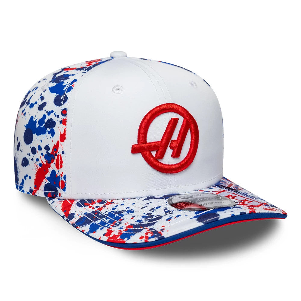 TGR Haas F1 Esteban Ocon Silverstone Race Special White New Era 9SEVENTY Stretch Snap Cap, three-quarter view showing the red embroidered Haas logo on the white front panel, with a multicoloured splatter design on the visor.