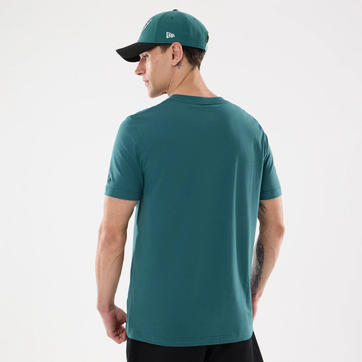 New Era Philadelphia Eagles NFL Training 2026 Dark Green T-Shirt, back view on a model styled with a matching cap, showing a plain dark green back with an embroidered logo on the left sleeve near the hem and short sleeves.