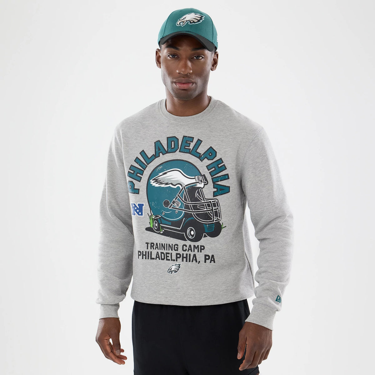 New Era Philadelphia Eagles NFL Training 2026 Grey Crew Neck Sweater, front view on a model wearing a matching cap, displaying Philadelphia lettering, an eagle graphic on a helmet, and NFL logo above Training Camp Philadelphia, PA.