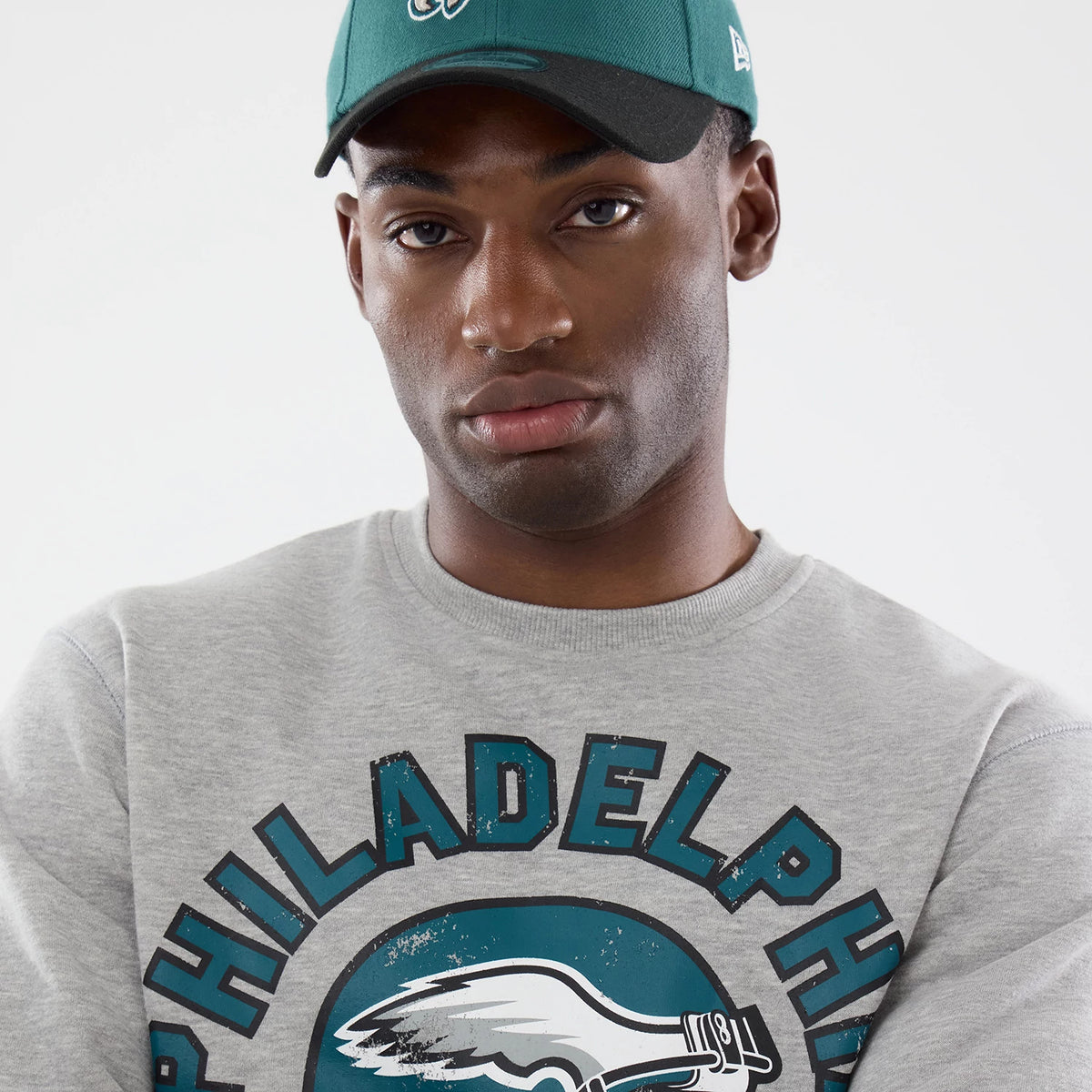 New Era Philadelphia Eagles NFL Training 2026 Grey Crew Neck Sweater, on-model front view featuring Philadelphia Eagles lettering and logo across the chest, paired with a green cap having the Eagles logo embroidered in white.