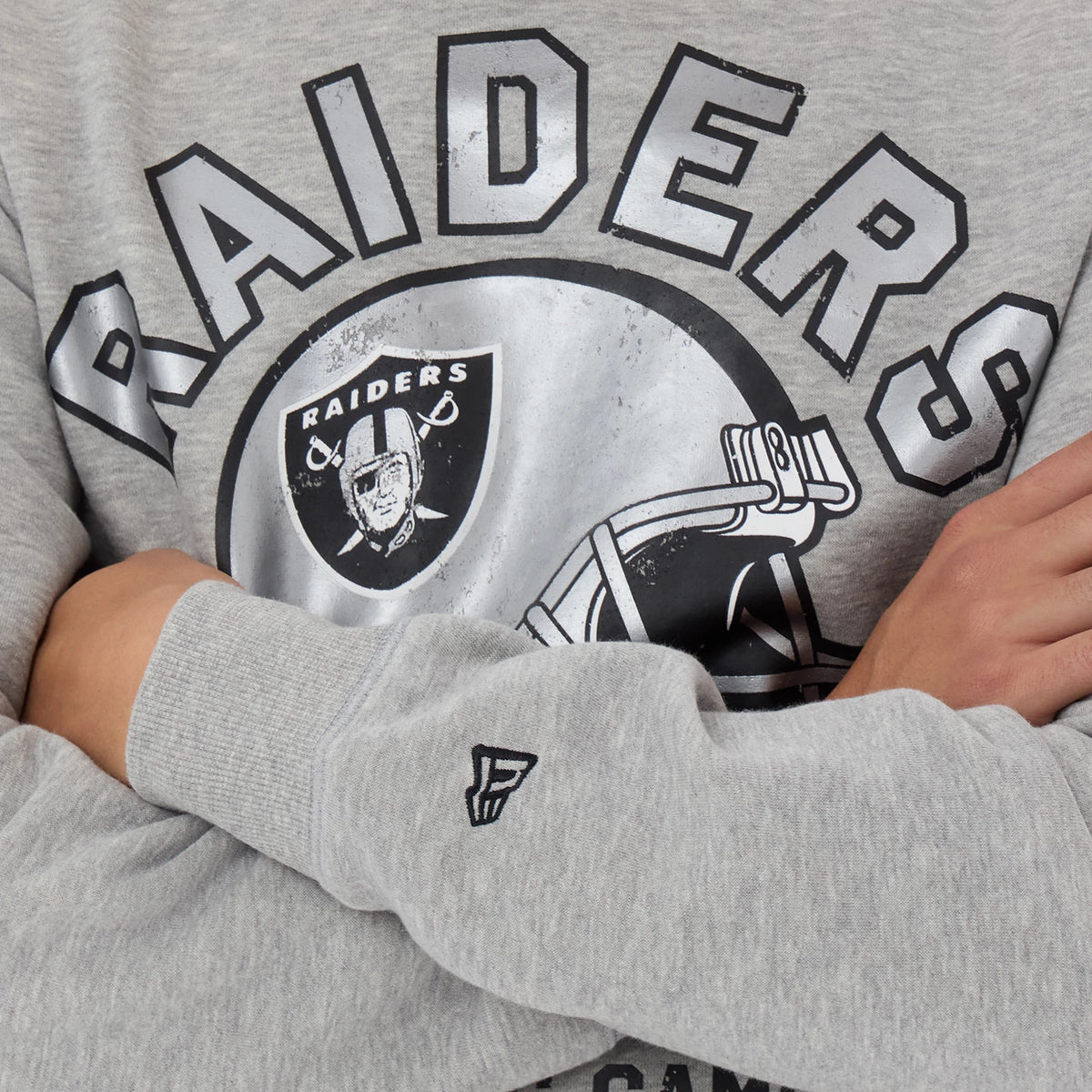 New Era Las Vegas Raiders NFL Training 2026 Grey Crew Neck Sweater, close-up view on a model, featuring the distressed black Raiders helmet graphic and lettering across the chest, with a black New Era logo embroidered on the grey sleeve cuff.