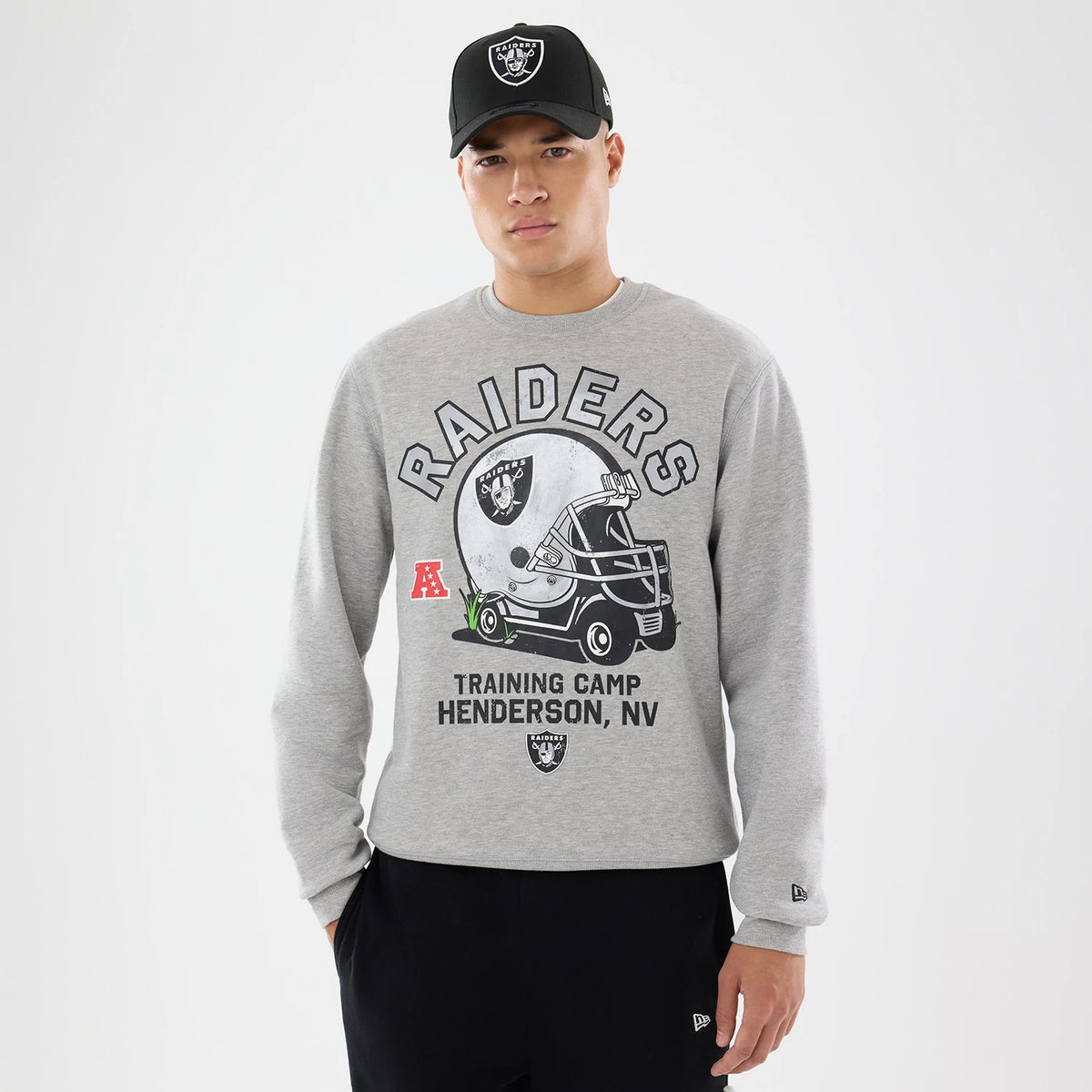 New Era Las Vegas Raiders NFL Training 2026 Grey Crew Neck Sweater, front view on a model wearing a black Raiders cap, featuring Raiders lettering and a helmet graphic on the chest, with AFC and New Era logos on the sleeves.