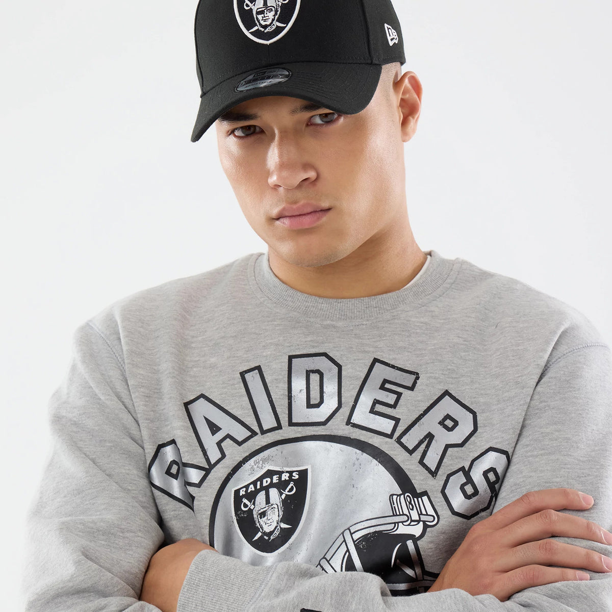 New Era Las Vegas Raiders NFL Training 2026 Grey Crew Neck Sweater, front view on a model with a black Raiders cap, showing the Raiders logo and lettering across the chest, with a ribbed collar and long sleeves.