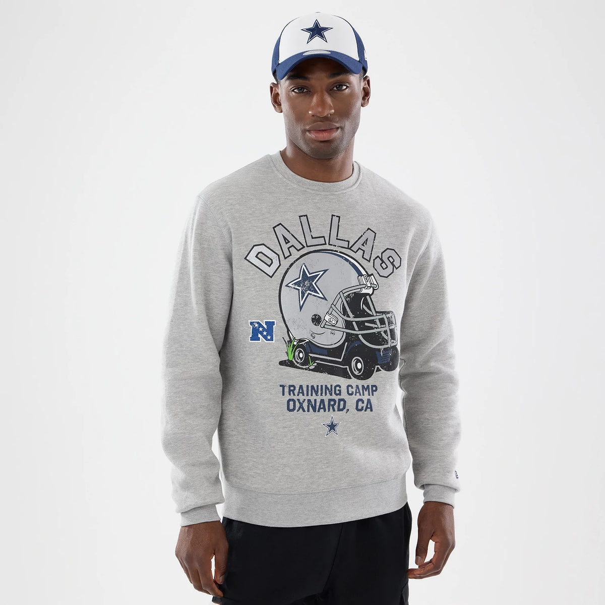 New Era Dallas Cowboys NFL Training 2026 Grey Crew Neck Sweater, front view on a model wearing a cap, featuring a large graphic of a helmet and Dallas text across the chest, with training camp details in blue underneath.