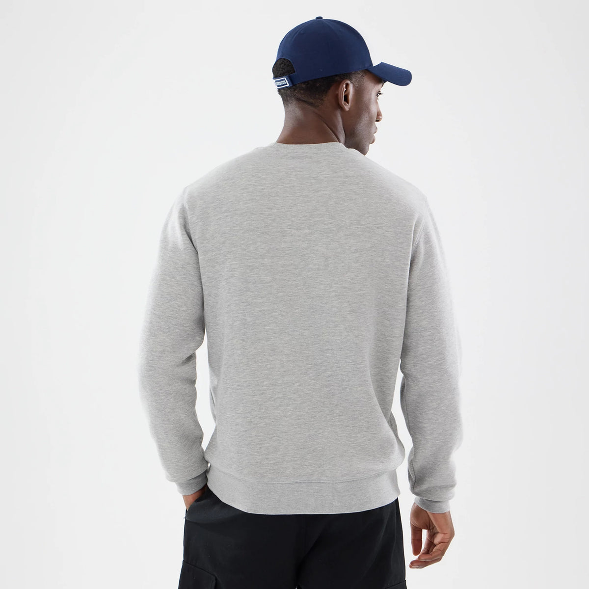 New Era Dallas Cowboys NFL Training 2026 Grey Crew Neck Sweater, rear view on a model with a navy cap, showing the plain light grey back panel of the sweater, ribbed cuffs, and a relaxed fit over black trousers.