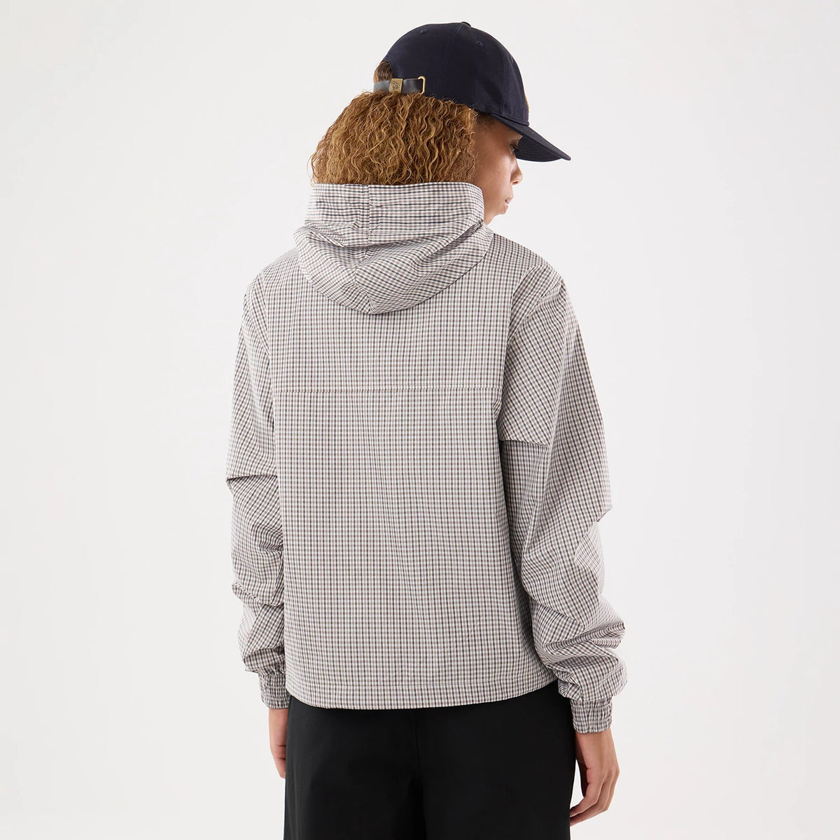 Womens New Era Check Beige Oversized Windbreaker Jacket, back view on-model showing the beige check pattern throughout, a hood resting on the shoulders, and relaxed long sleeves with elastic cuffs, paired with a dark cap and trousers.