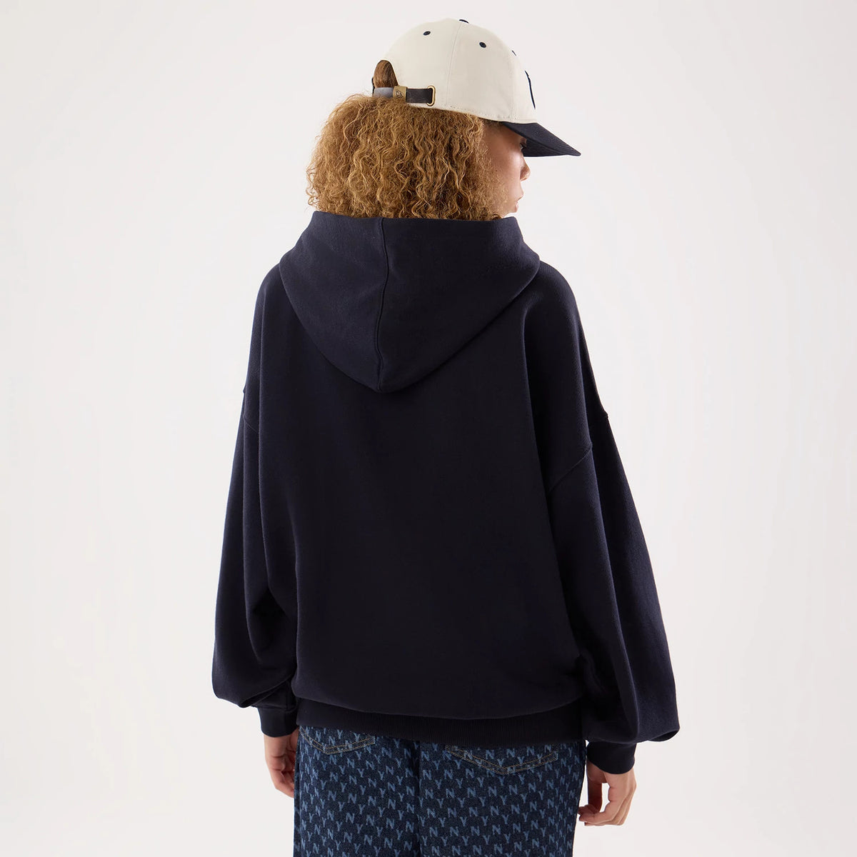 New Era New York Yankees Womens MLB Navy Oversized Pullover Hoodie, back view on a model, showcasing the loose-fitting navy hooded sweatshirt with relaxed sleeves and hem, styled with patterned trousers and a light-coloured cap with eyelets.