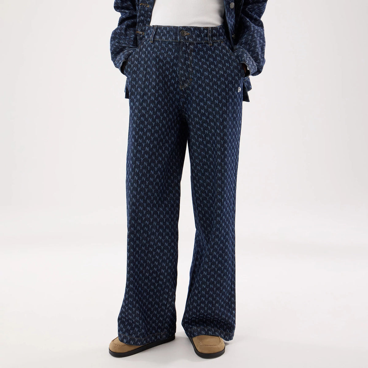 New Era New York Yankees Womens MLB Denim Navy Wide Leg Trousers, front view on a model wearing a matching jacket, showing an all-over repeating Yankees logo pattern, belt loops, and wide leg hems reaching down to beige shoes with black soles.