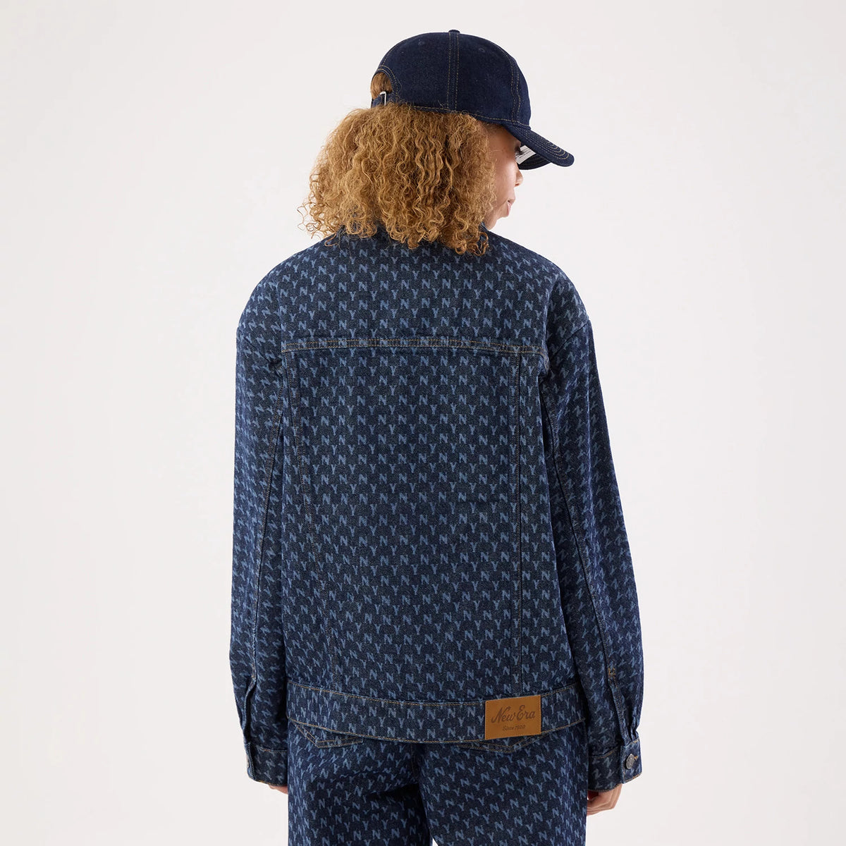 New Era New York Yankees Womens MLB Denim Navy Jacket, back view on a model styled with a matching cap, displaying a repeating NY pattern across the dark navy fabric and a tan brown patch with an embossed New Era logo at the lower hem.