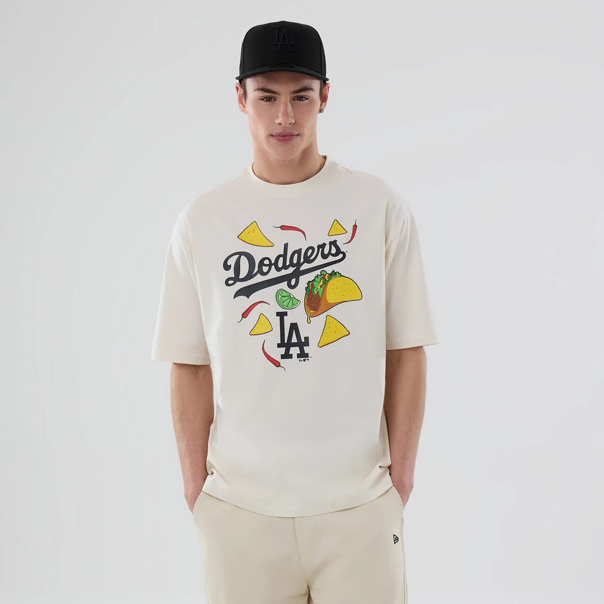 New Era LA Dodgers MLB Food Graphic Open White Oversized T-Shirt, front view on-model showcasing Dodgers script with graphic elements including tacos, chilli peppers, and nachos on a white backdrop, LA logo below, paired with a black cap.