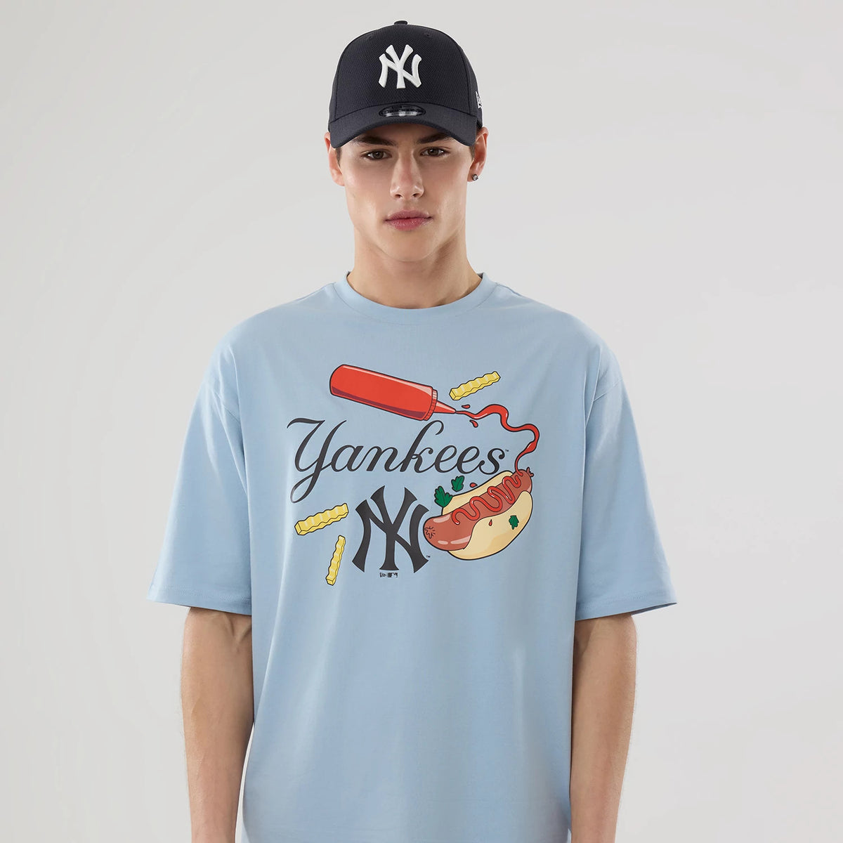 New Era New York Yankees MLB Food Graphic Pastel Blue Oversized T-Shirt, front view on a model featuring a ketchup bottle and hot dog graphic with fries, complemented by embroidered Yankees lettering.