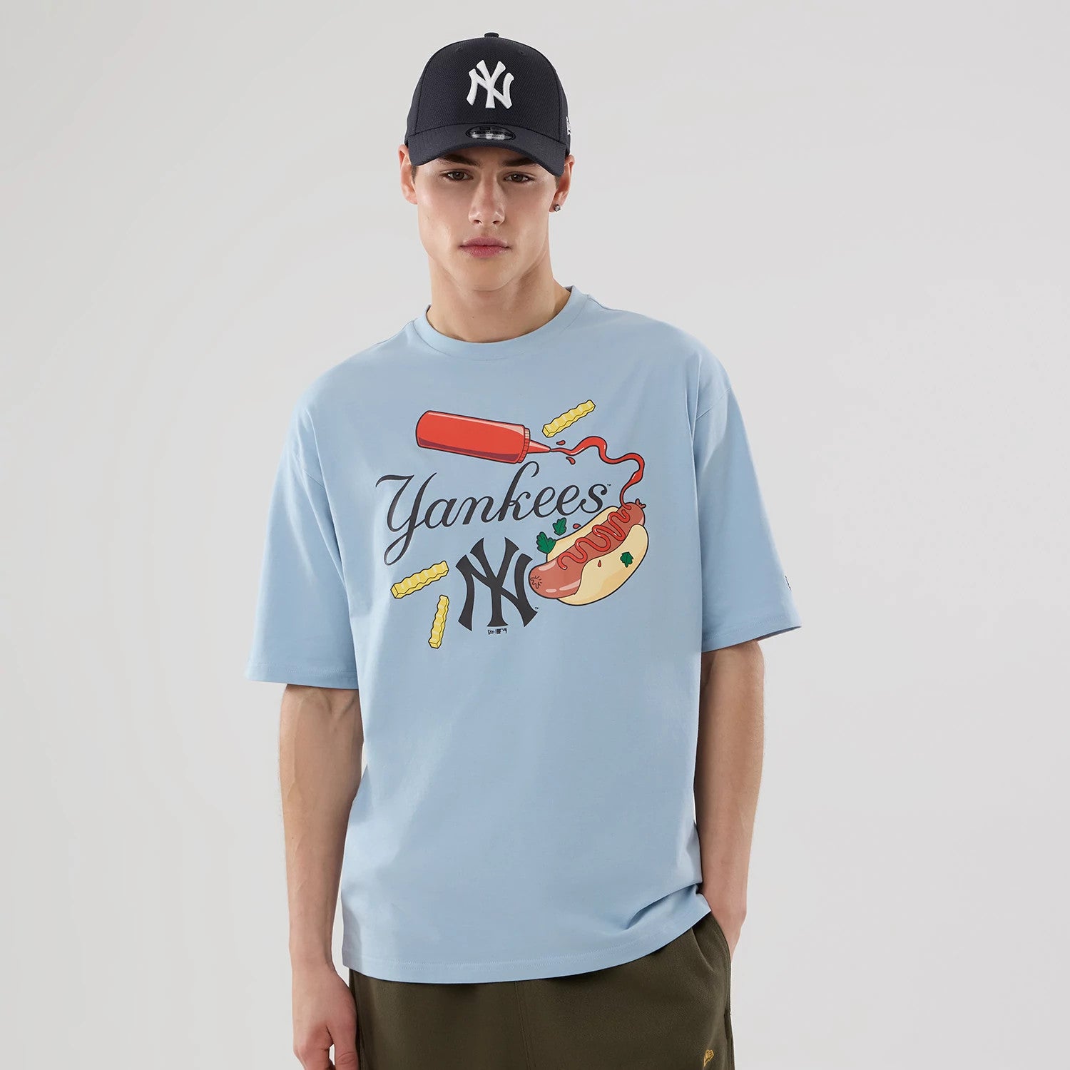 New Era New York Yankees MLB Food Graphic Pastel Blue Oversized T-Shirt, front view on a model wearing a navy cap, with a playful graphic of a hot dog, ketchup bottle, fries and the Yankees logo beneath styled script on the pastel blue fabric.