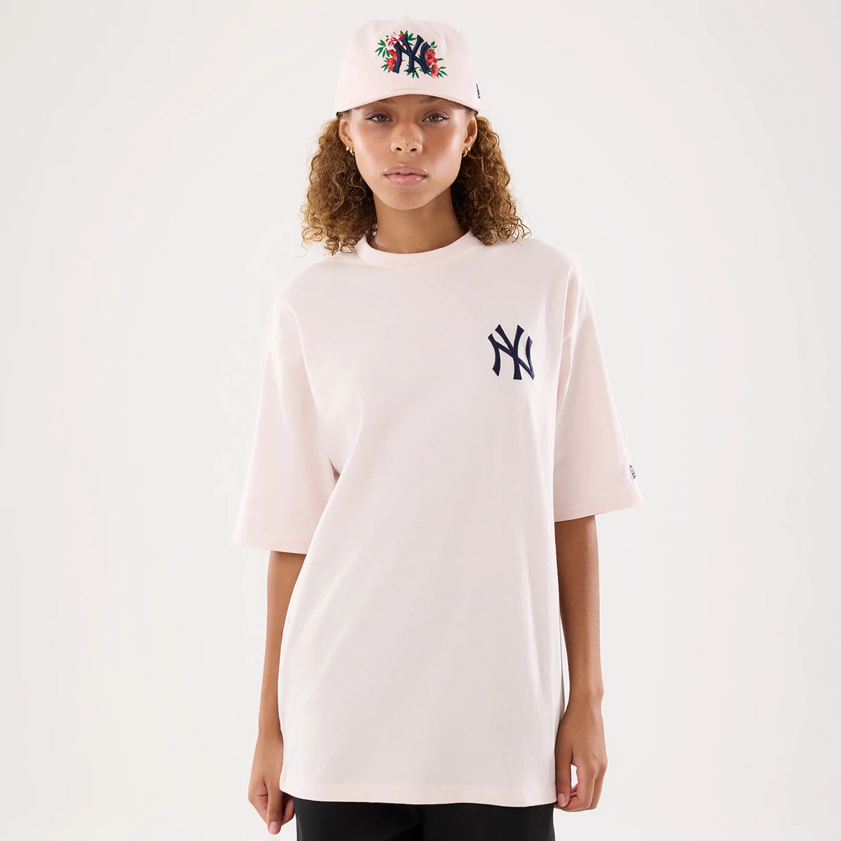 New Era New York Yankees Womens MLB Graphic Pastel Pink T-Shirt, front view on-model showing the oversized pastel pink T-shirt with a navy embroidered Yankees logo on the left chest, styled with a matching cap featuring floral embroidery.