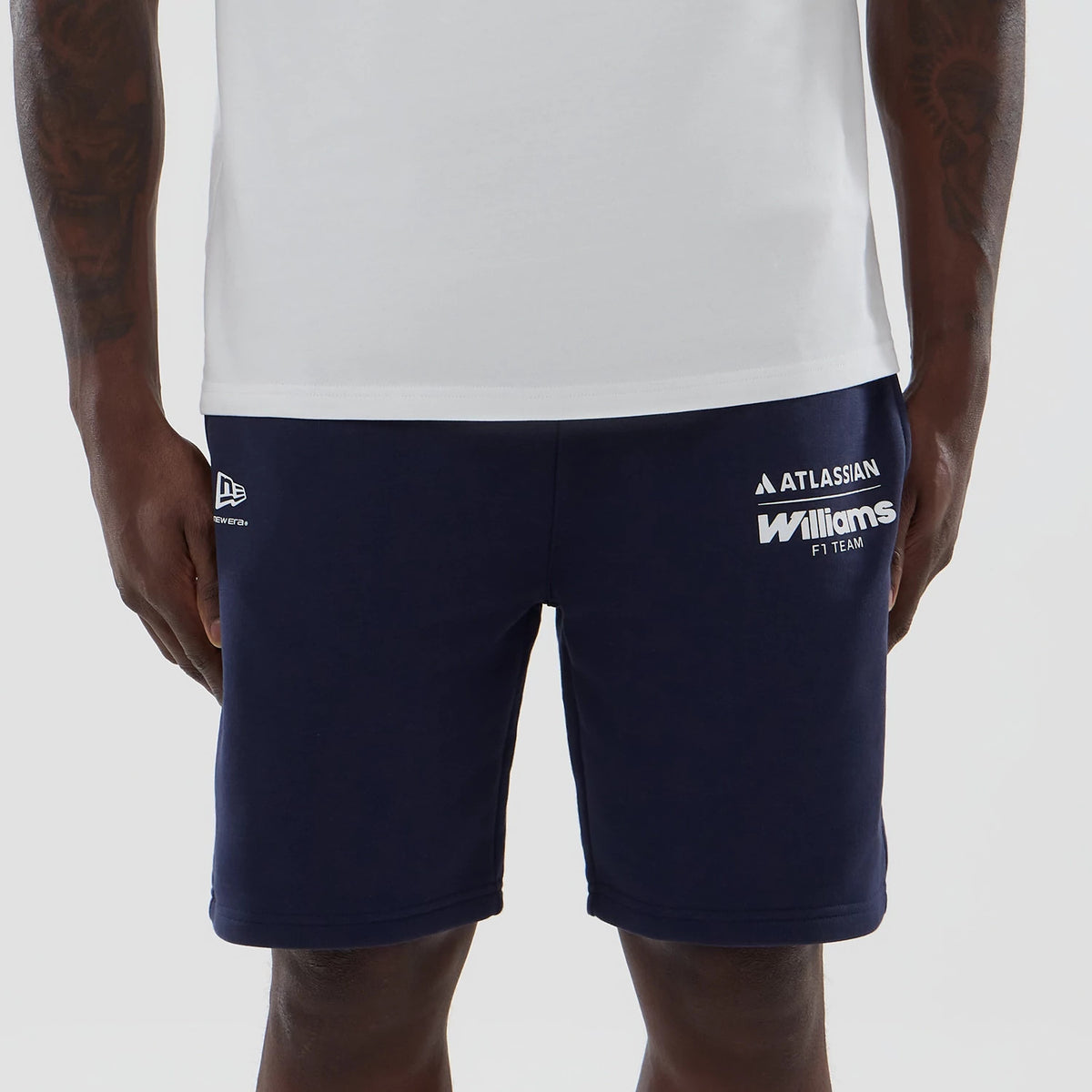 New Era Williams Navy Shorts, front view on a model showing the navy fabric with white printed Williams F1 Team and Atlassian logos on the right leg, and an embroidered New Era logo near the left hem, styled with a plain white top.