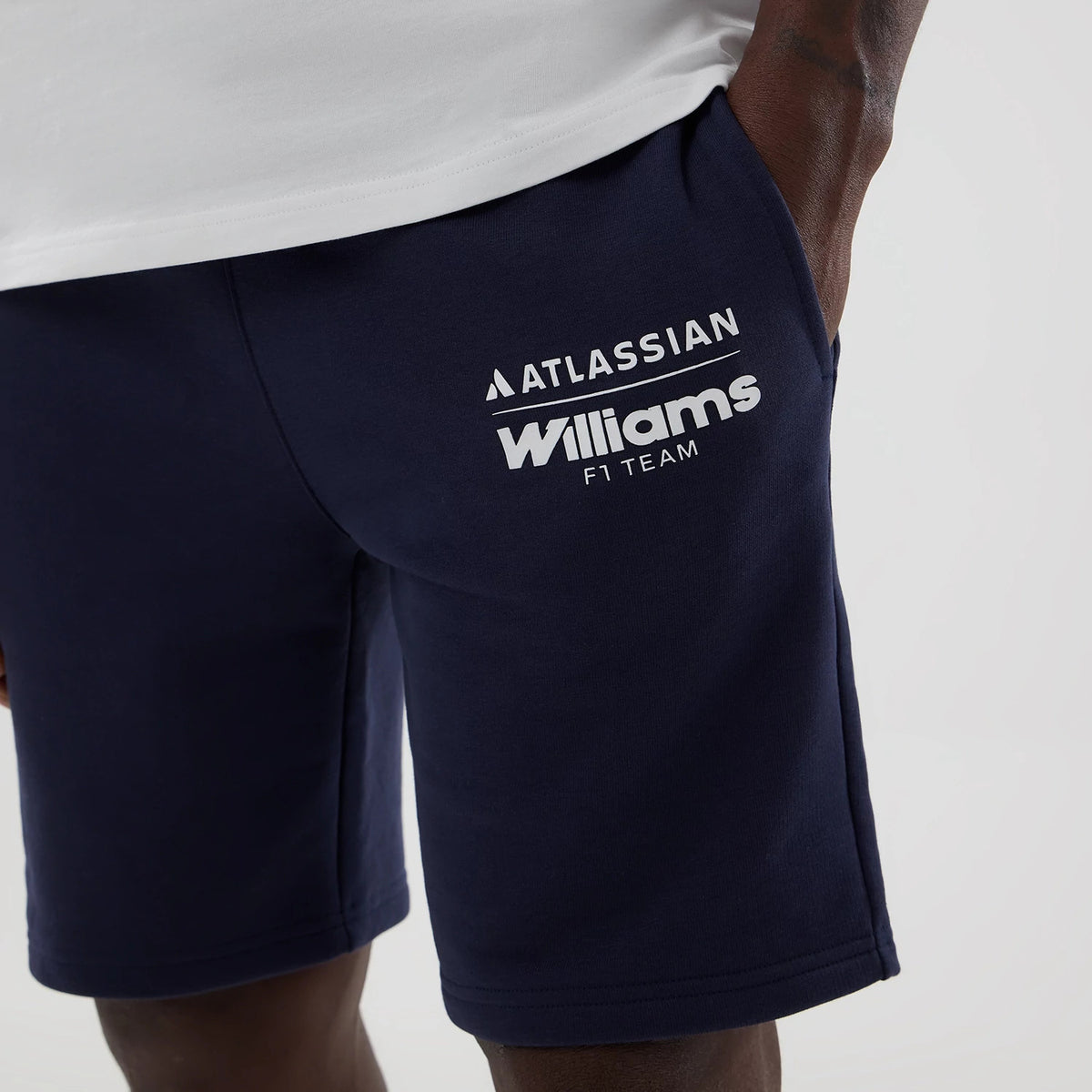 New Era Williams Navy Shorts, front view showcasing the navy blue design worn with a white top, featuring a printed white Atlassian and Williams F1 Team logo on the left thigh, with the shorts' pocket opened slightly by the hand.