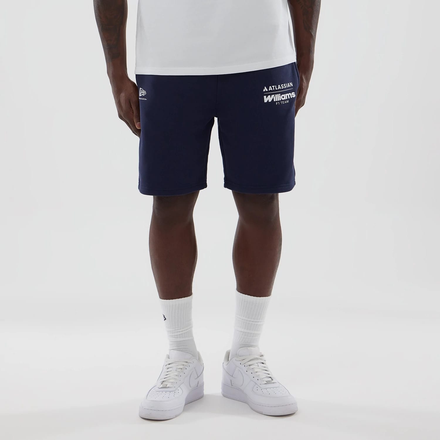 New Era Williams Navy Shorts, front view on-model, showcasing the navy colour with white printed team logos on the left side, the New Era logo embroidered on the opposite side, paired with a plain white t-shirt and matching white trainers.