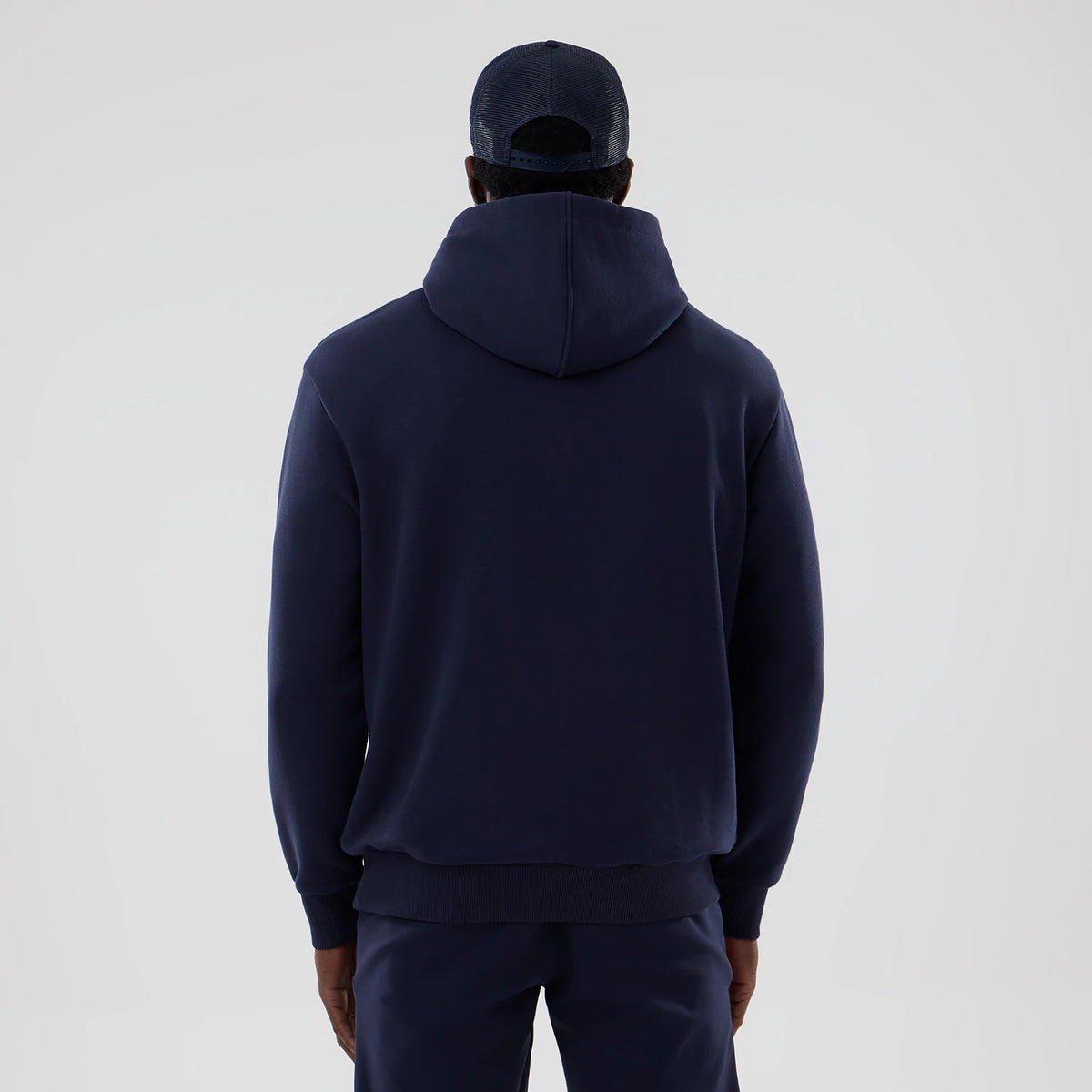 New Era Williams Navy Oversized Pullover Hoodie, back view on a model showing the hoodie’s relaxed fit with ribbed cuffs and hem, a spacious hood resting on the back, and paired with navy trousers and a dark cap.
