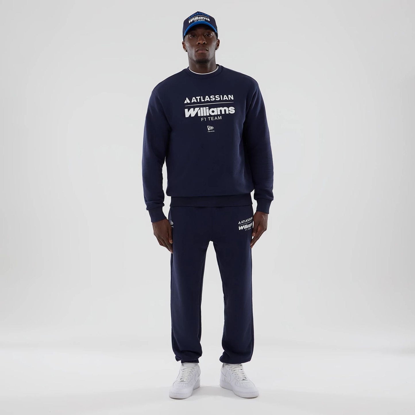 New Era Williams Navy Joggers, on-model view with a matching navy sweatshirt featuring white branding on the top left leg, elasticated cuffs, and paired with white trainers for a complete uniform look.