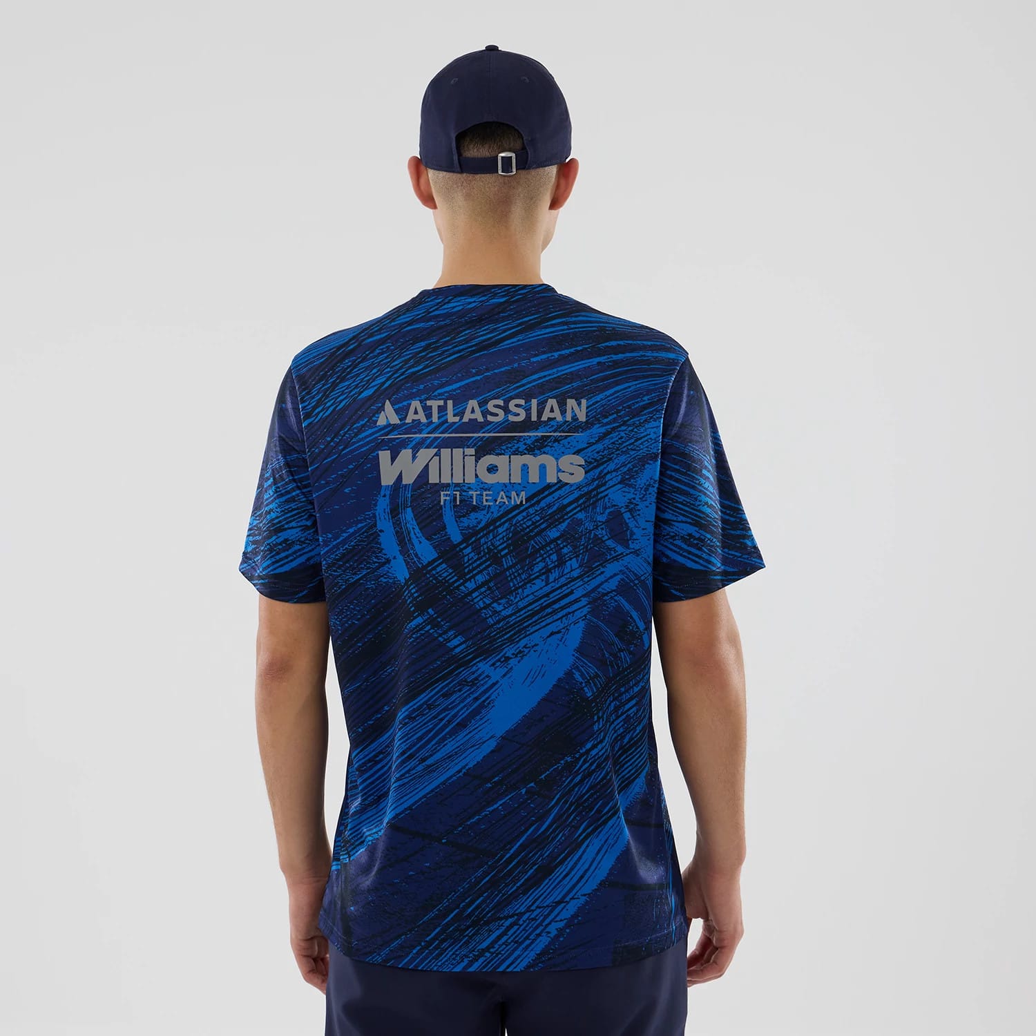 New Era Williams Navy Active T-Shirt, back view on a model with a matching navy cap, featuring Atlassian and Williams F1 Team lettering printed across the shoulders against a abstract navy and blue swirl pattern on the fabric.