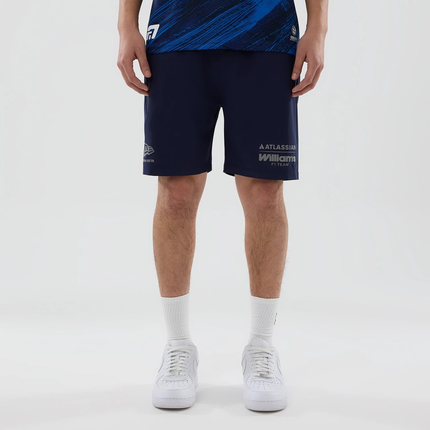 New Era Williams Navy Training Shorts, front view on-model showing printed Williams F1 Team logo on the lower right leg, embroidered New Era logo on the lower left leg, and a navy body with a relaxed fit styled with white trainers and socks.