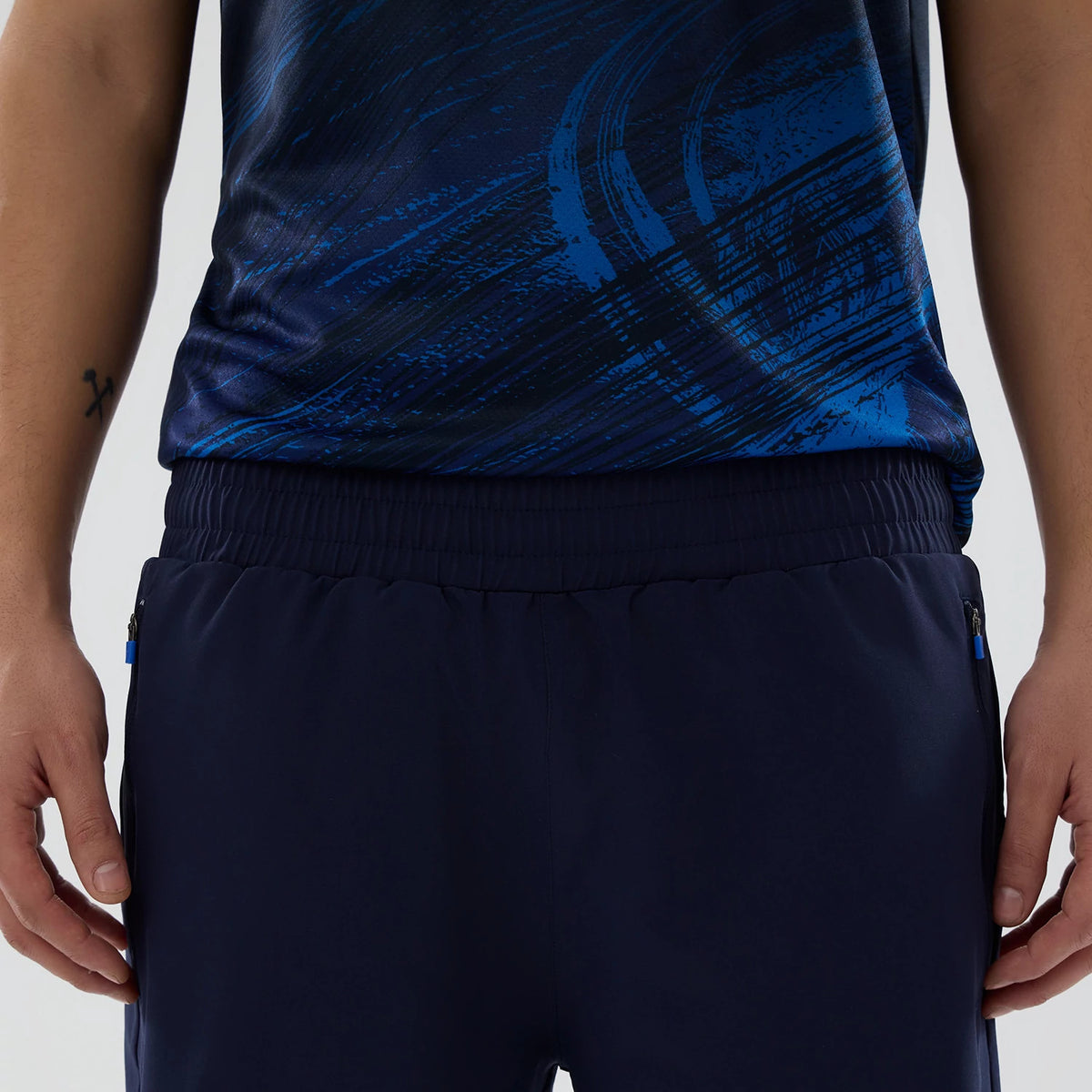 New Era Williams Navy Training Shorts, front view showing the elastic waistband and two side zipped pockets with blue zip pulls, styled on a model with a dark blue patterned top complementing the navy shorts.