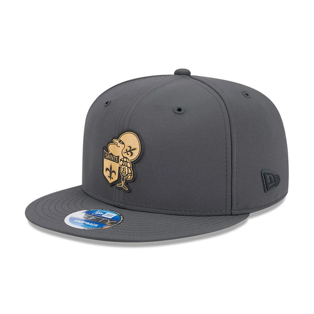 Saints NFL Sideline 2026 Charcoal New Era 9FIFTY Snapback Adjustable Cap, front view showing a gold embroidered character and Saints logo on the front panel, a flat visor with a 9FIFTY size sticker, and the New Era flag logo on the left side.