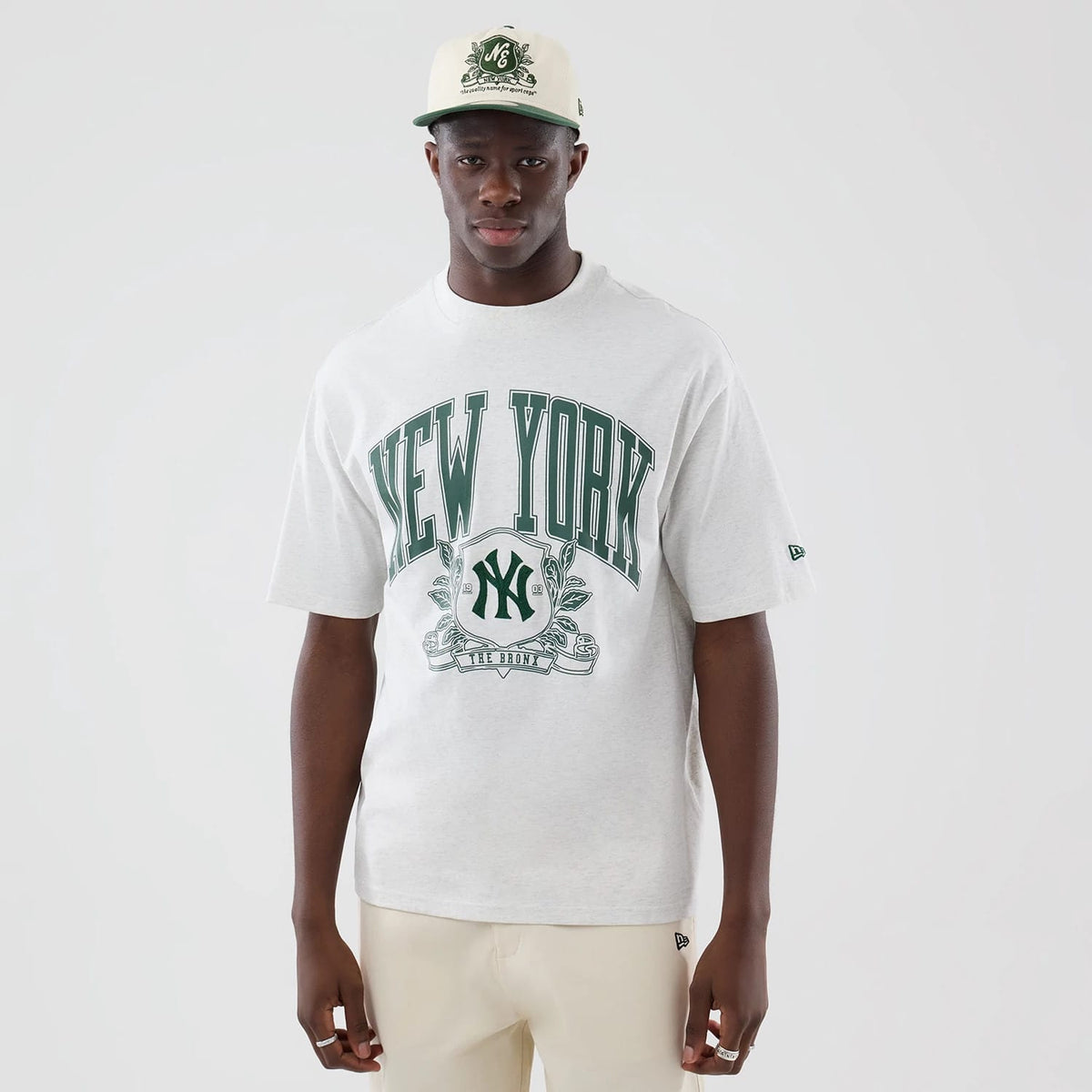 New Era New York Yankees MLB Arch Graphic Pastel Grey Oversized T-Shirt, front view on a model paired with a matching cap, showing large green New York lettering across the chest, an embroidered New Era logo on the sleeve and a central logo graphic.
