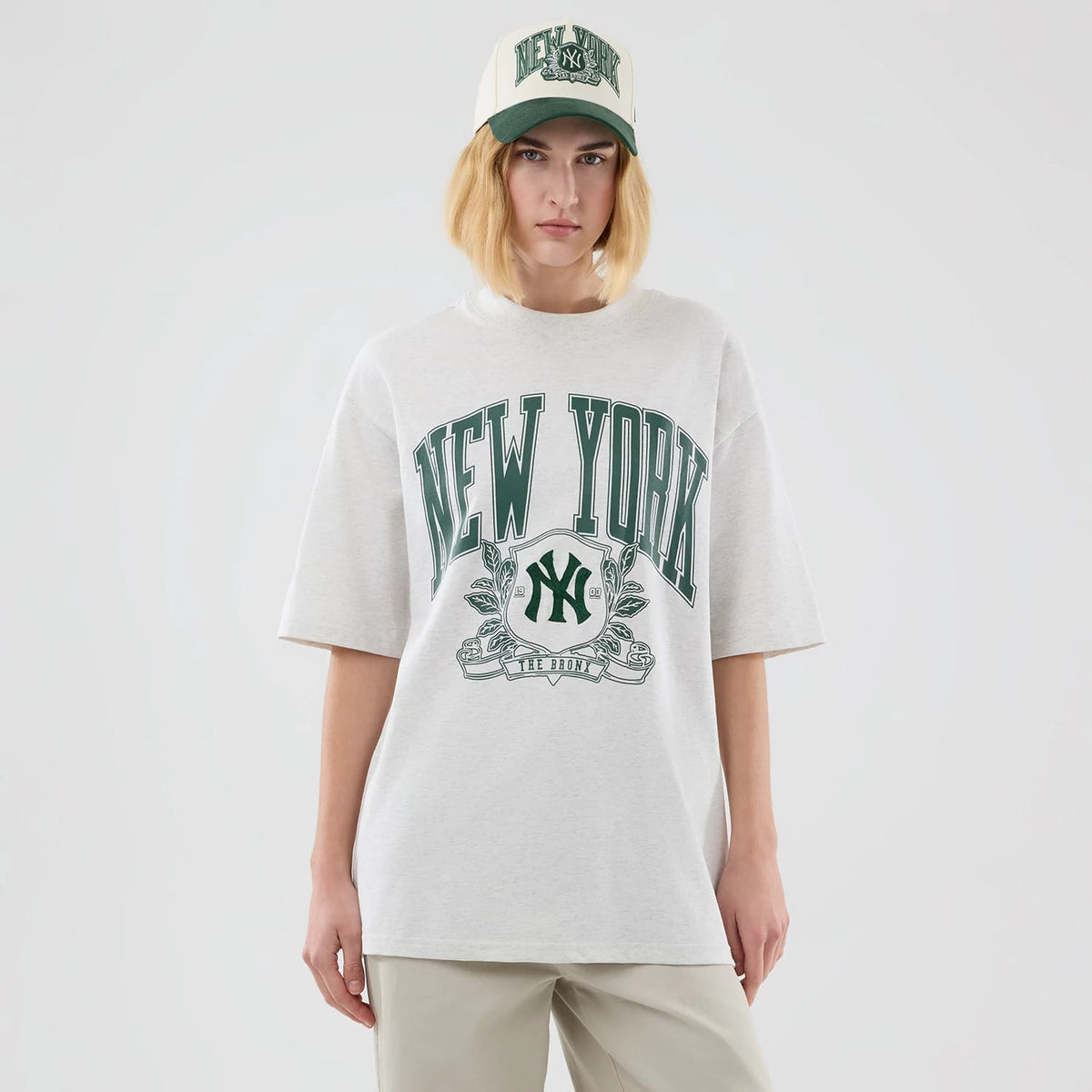 New Era New York Yankees MLB Arch Graphic Pastel Grey Oversized T-Shirt, front view on a model with matching cap, featuring large green New York lettering across the chest, an embroidered Yankees logo below it, and short sleeves.