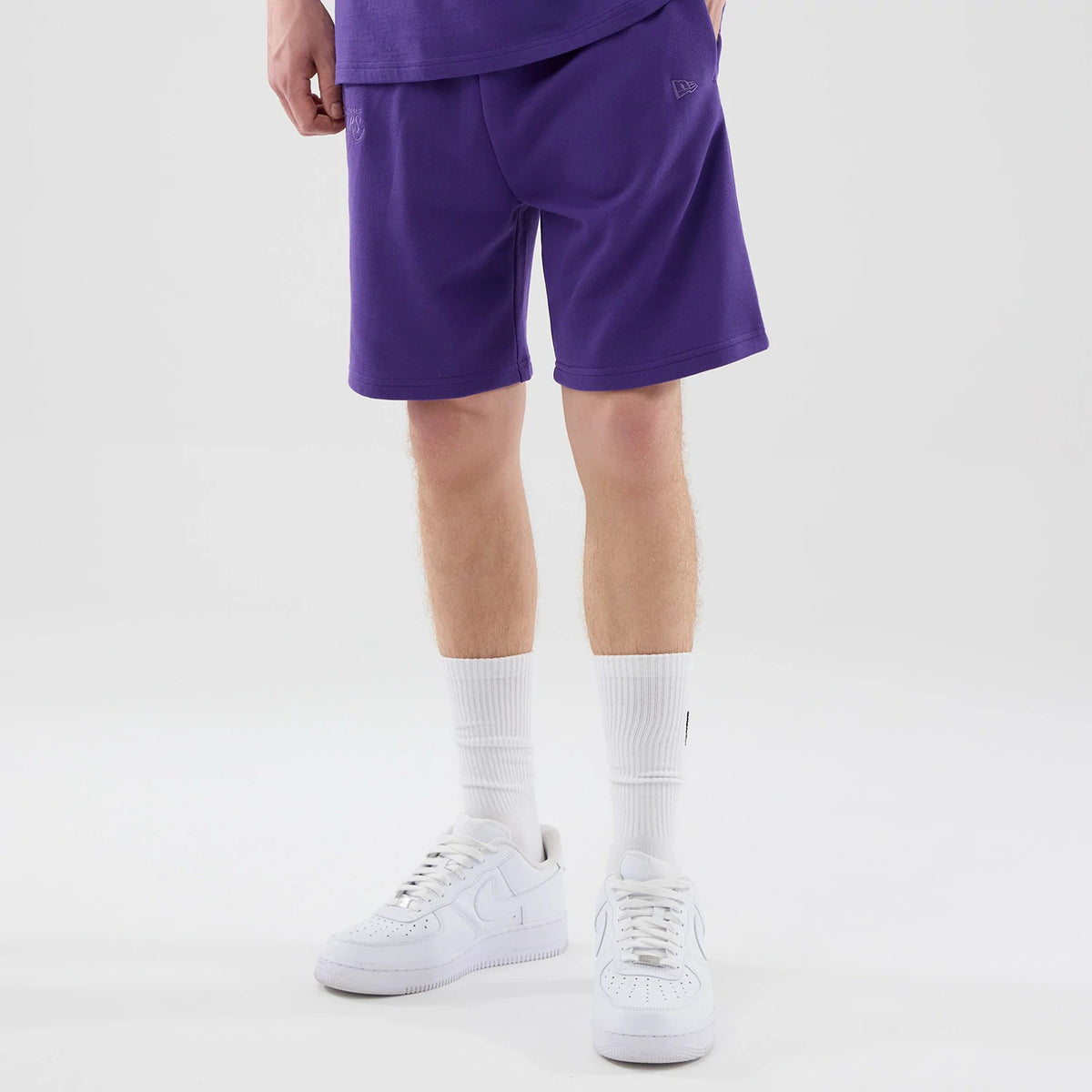 New Era LA Lakers NBA League Essential Purple Oversized Shorts, front view on a model styled with white trainers and socks, showing a relaxed fit with an embroidered New Era logo near the pocket and a plain hem along the knee-length silhouette.